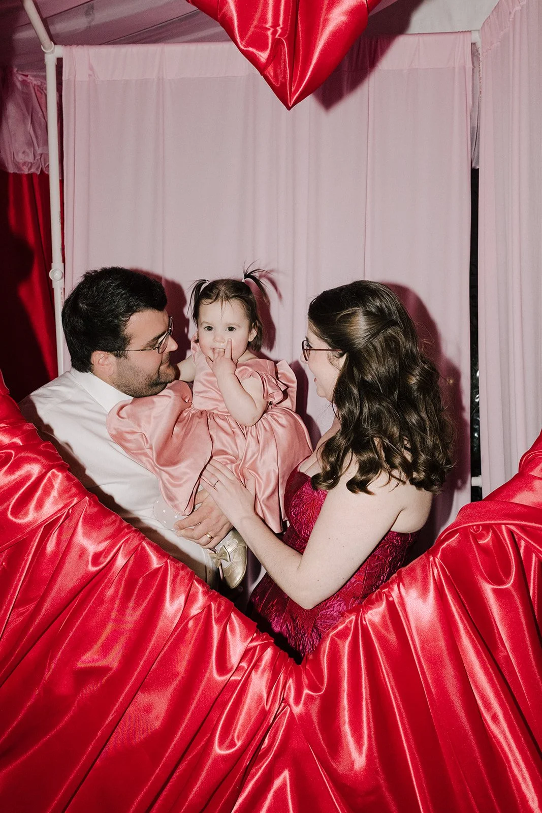 A family of three, a man, woman, and a young girl, inside a heart-shaped photo booth with pink curtains. The man is holding the girl, who is wearing a pink dress, while the woman in a red dress looks at them.