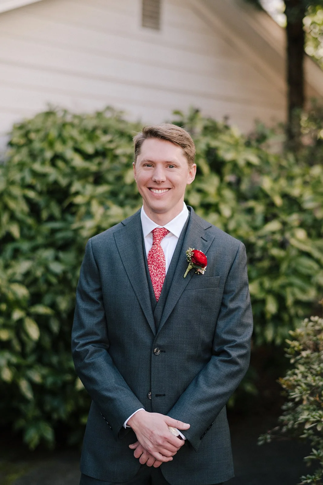 A man in a dark gray suit, white shirt, and red patterned tie stands outdoors, smiling, with a red boutonniere on his lapel and green shrubbery in the background.
