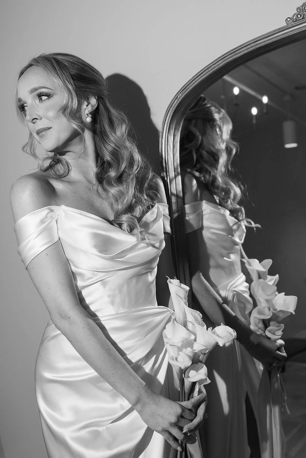 A woman in a satin dress holding flowers and looking to the side, with her reflection in an ornate mirror beside her.