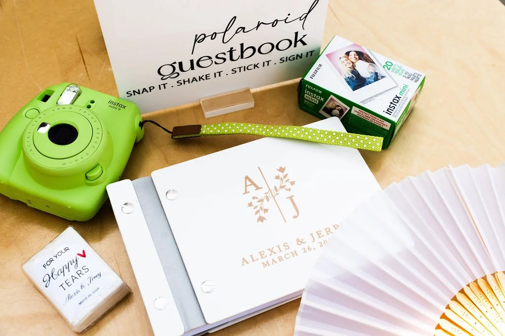 Wedding guestbook, instant camera, film, a small box of tissues, a hand fan, and a card with the names Alexis and Jerdy, and the date March 26, 2023, on a wooden surface.