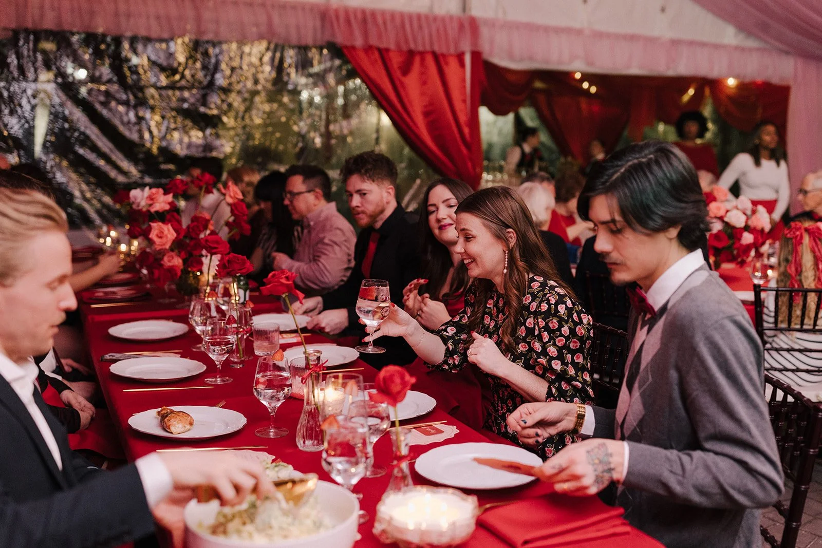 People gathered around a long table decorated with red roses for a formal event, with some people enjoying food and drinks, in a festive tent setting with curtains and hanging lights.