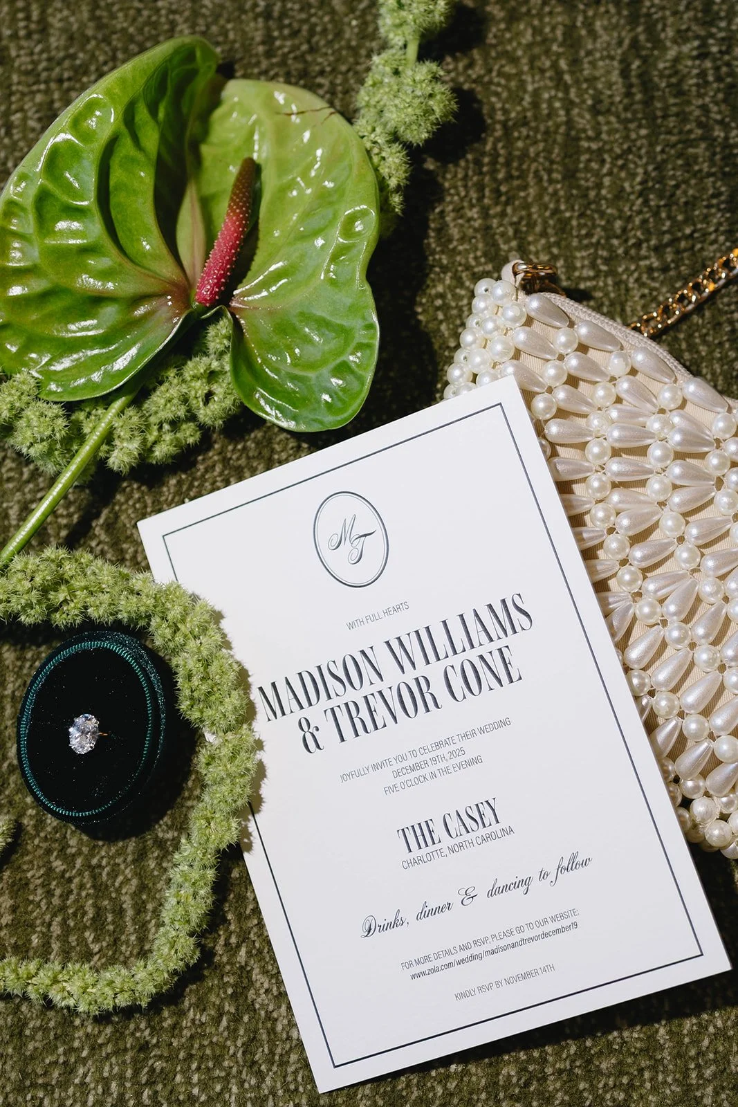 Wedding invitation surrounded by a pearl clutch, an engagement ring in a velvet box, a green floral arrangement, and decorative greenery.