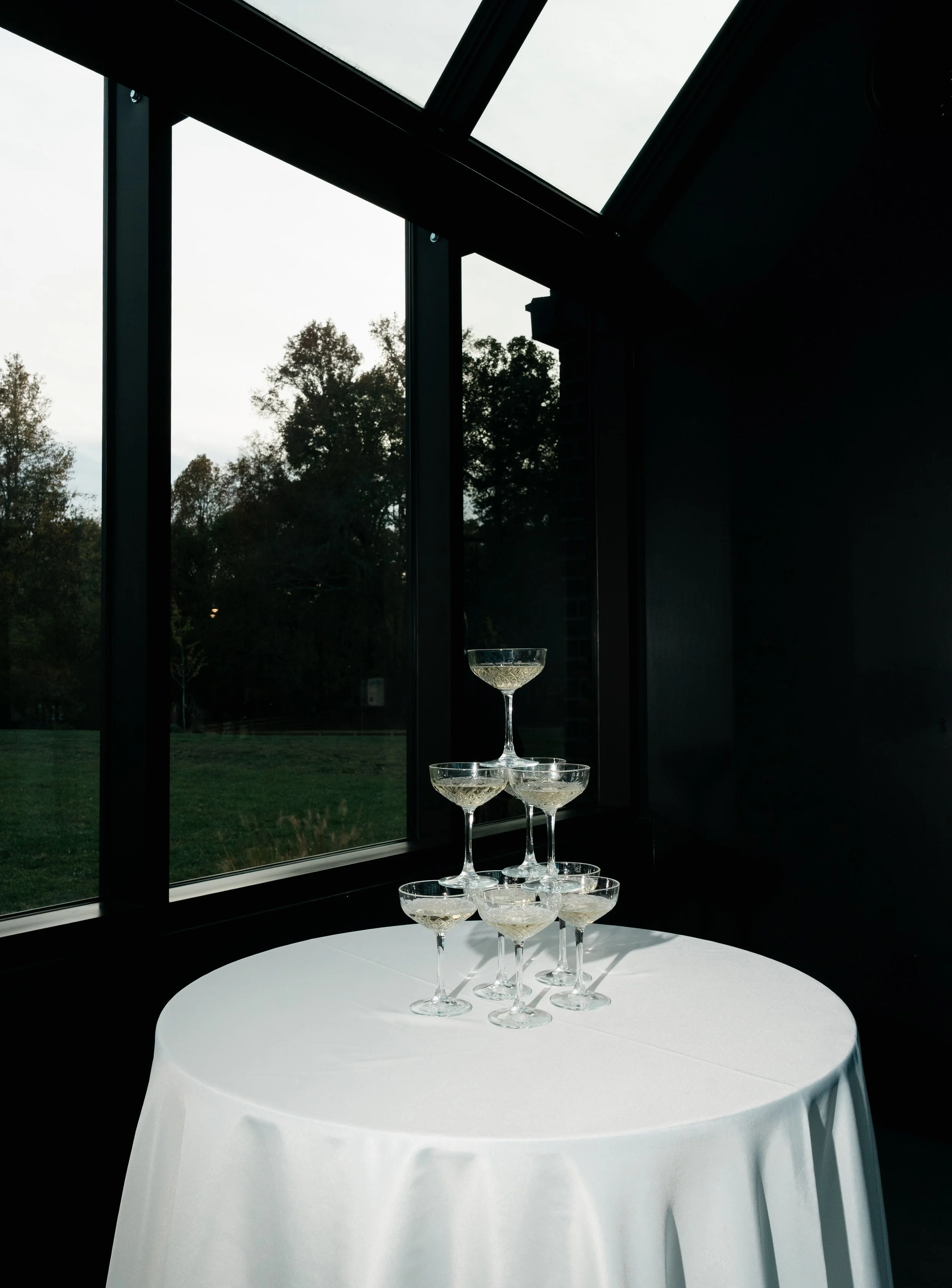 A pyramid of champagne glasses on a white round table near a large window showing trees outside on an overcast day.