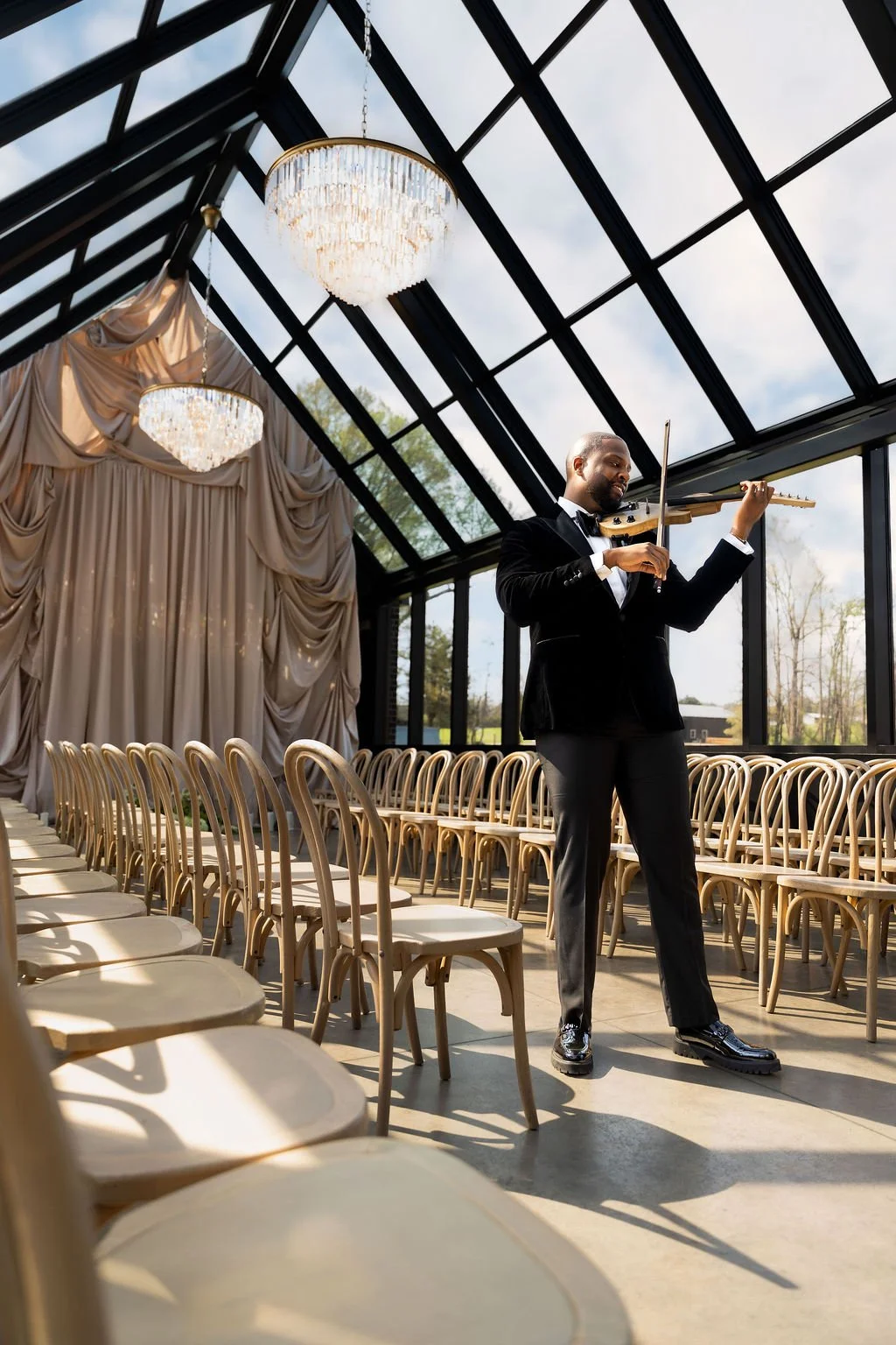 A man in a black tuxedo playing a violin in a bright, glass-walled room with chandeliers and empty chairs.