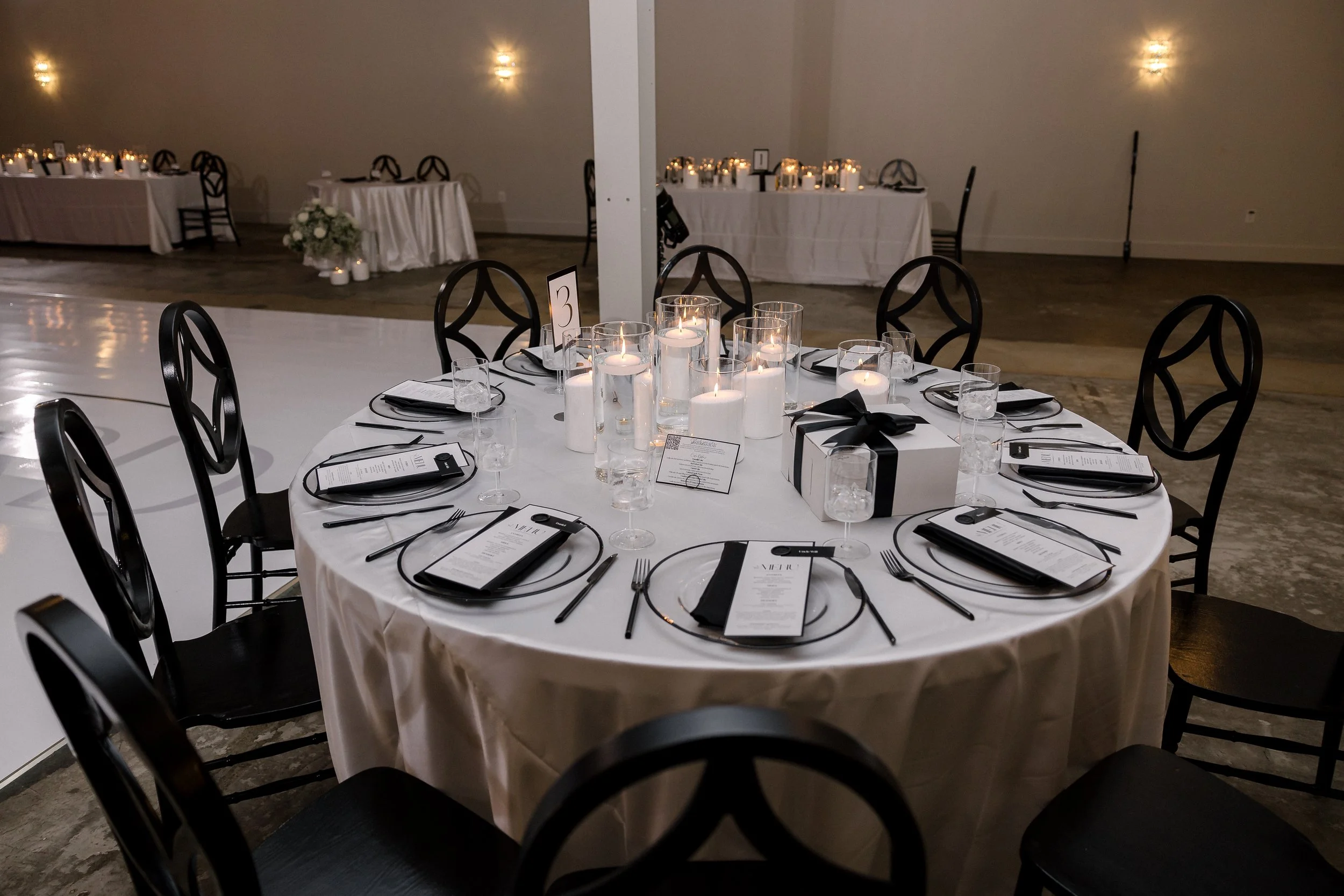 Round banquet table with black chairs, white tablecloth, black napkins, and place settings for a formal event, decorated with tall white candles and a small darker container with a menu card. In the background, there are additional tables with candle