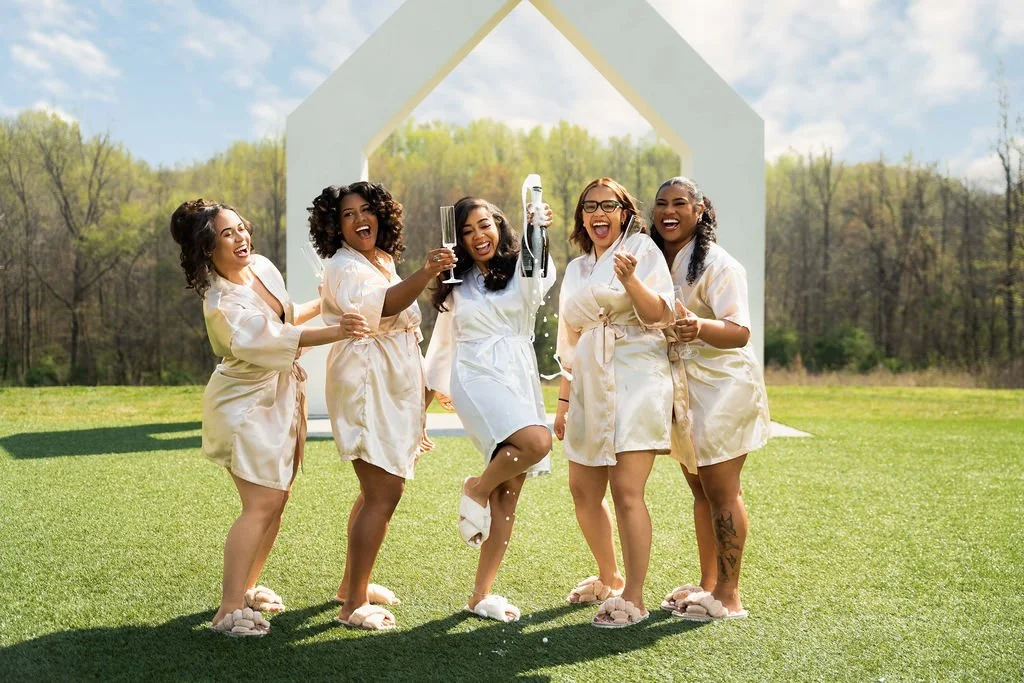 Five women in matching robes celebrating outdoors on a sunny day, one holding a champagne bottle, under a large white arch with trees in the background.