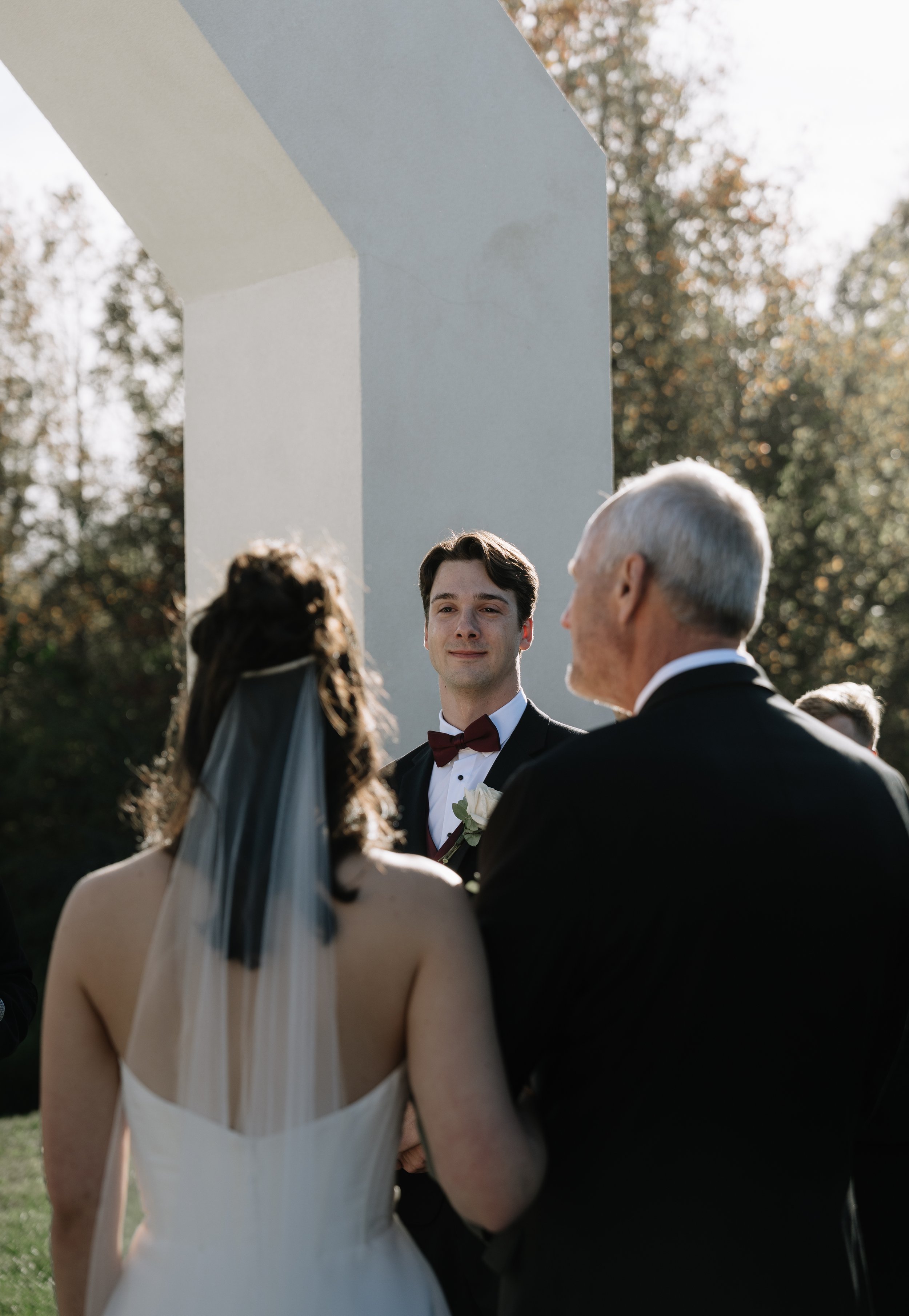 A wedding ceremony outdoors with a bride, groom, and officiant standing near a white monument under trees with sunlight filtering through.
