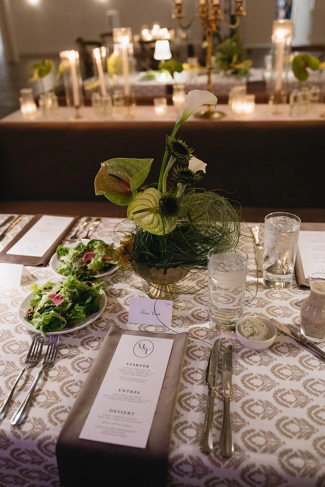Elegant dining table with a floral centerpiece, salad, glasses of water, cutlery, and a menu card, set in a dimly lit decorated room with candles and a chandelier.