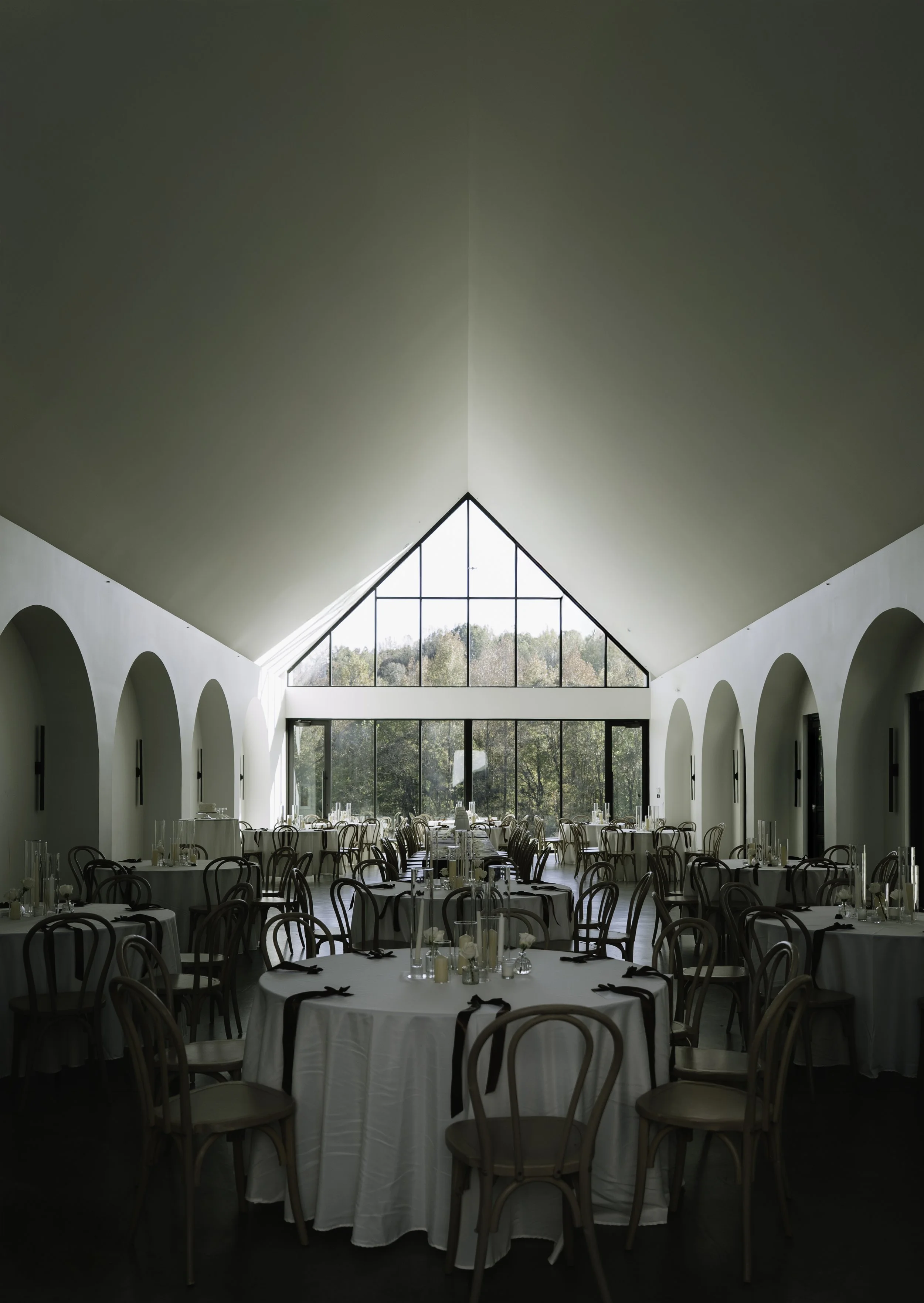 Elegant banquet hall with round tables covered in white tablecloths, decorated with candles and glassware, surrounded by wooden chairs, large window with triangular design letting in natural light, and trees visible outside.