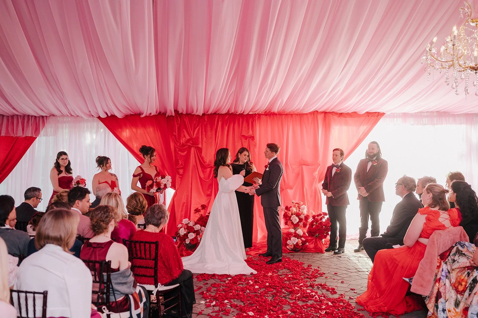 A wedding ceremony with a bride and groom exchanging rings, surrounded by bridesmaids and groomsmen, in a venue decorated with pink and red drapery and flowers, with guests seated watching.