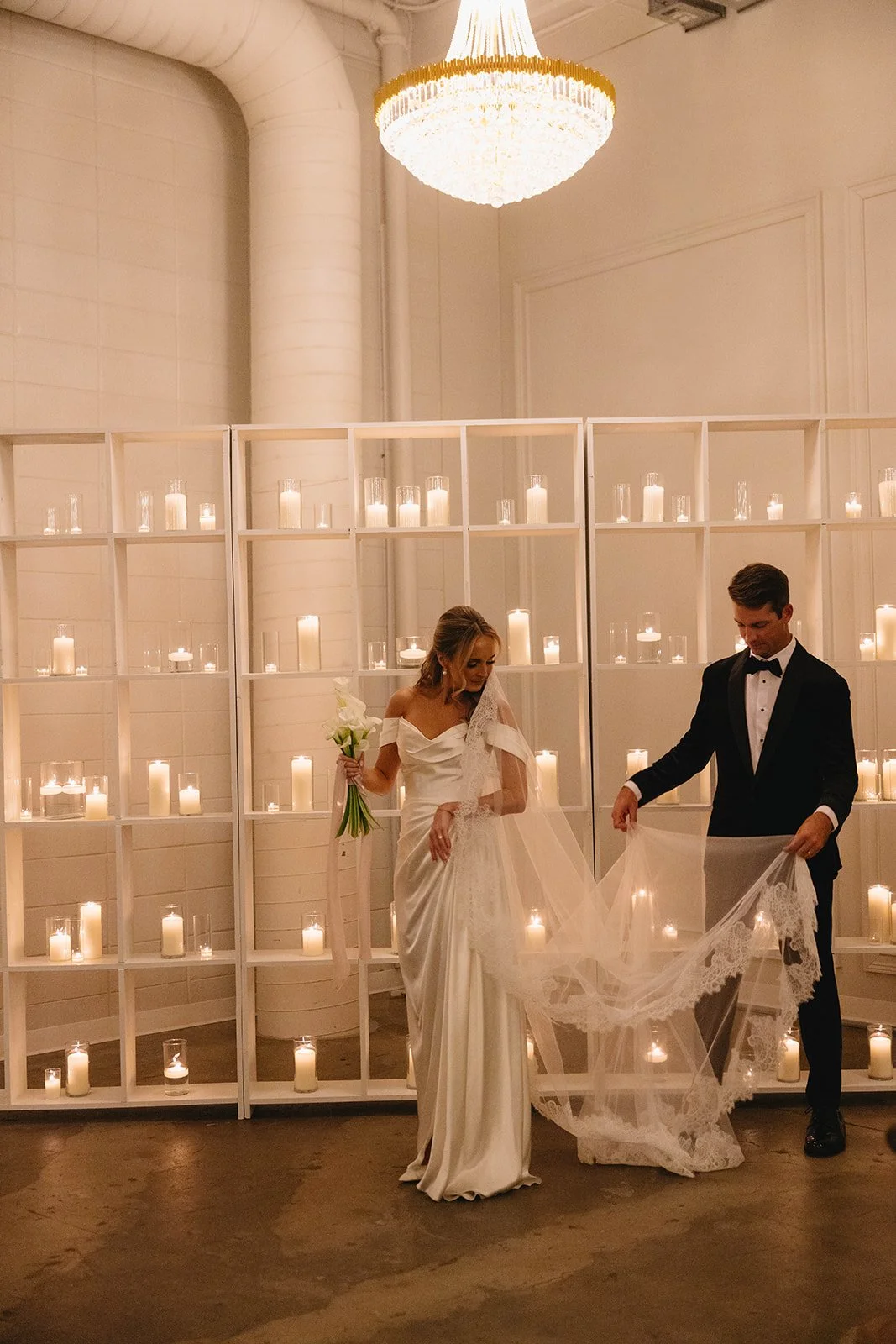Bride and groom standing in front of a wall of lit candles, with the bride holding a bouquet of white flowers, as they prepare for their wedding ceremony.