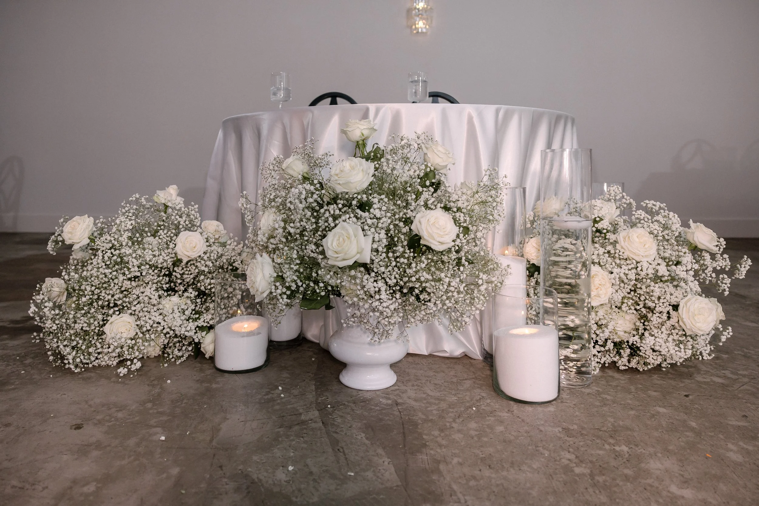 A decorative floral arrangement with white roses and baby's breath, surrounded by white candles and glass vases filled with water, set on a concrete floor against a neutral wall.