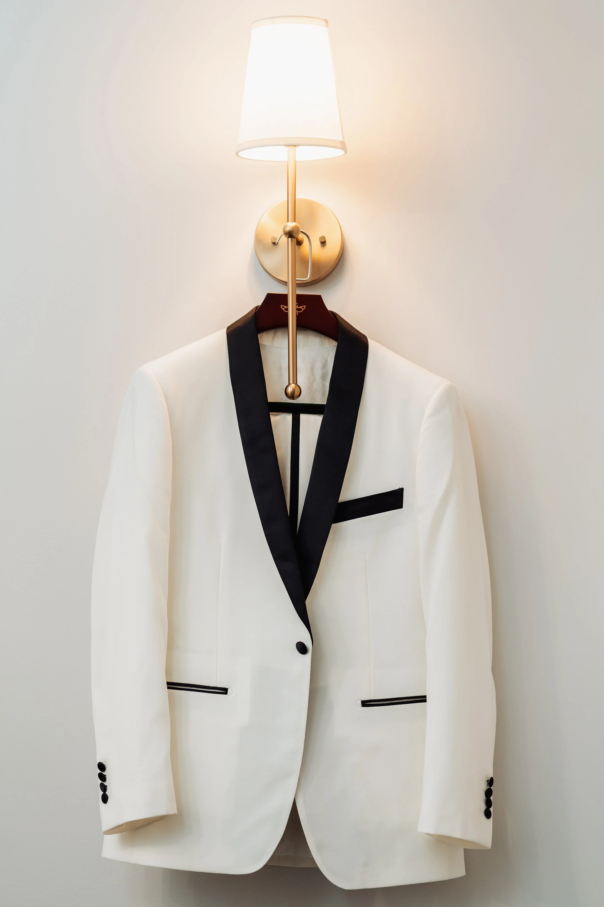 White tuxedo jacket with black trim on lapels, pockets, and buttons hanging on a hanger against a plain wall, with a wall-mounted lamp above.