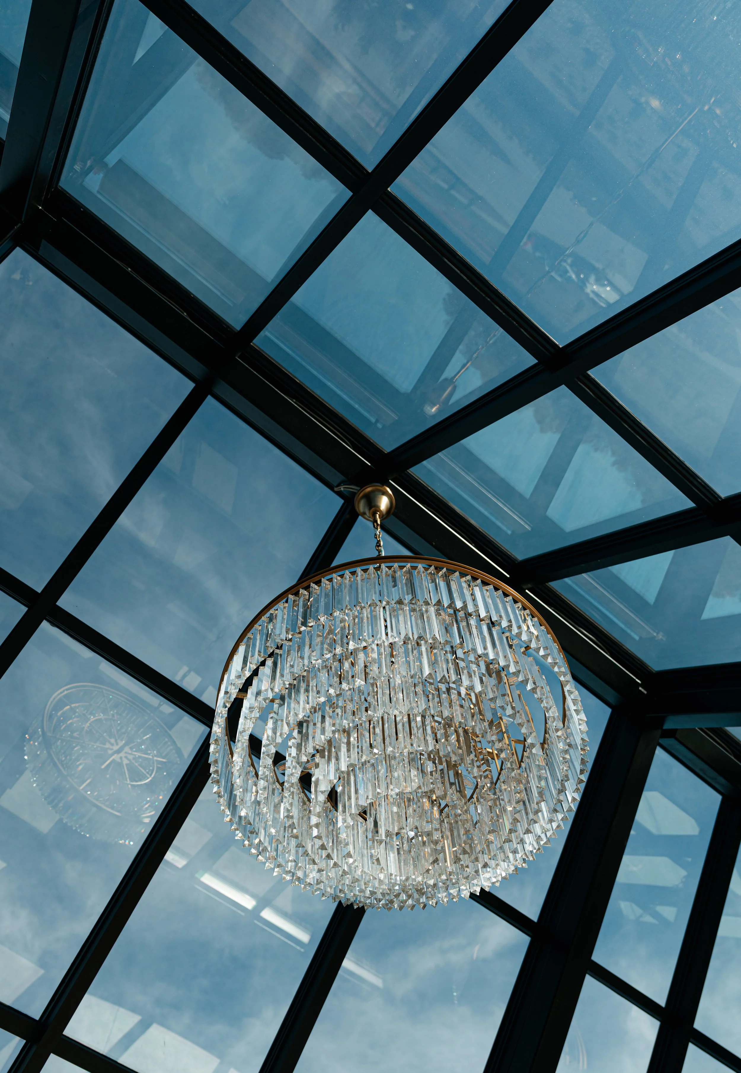 Chandelier hanging inside a glass ceiling structure, with outdoor sky and clouds visible through the glass panels.