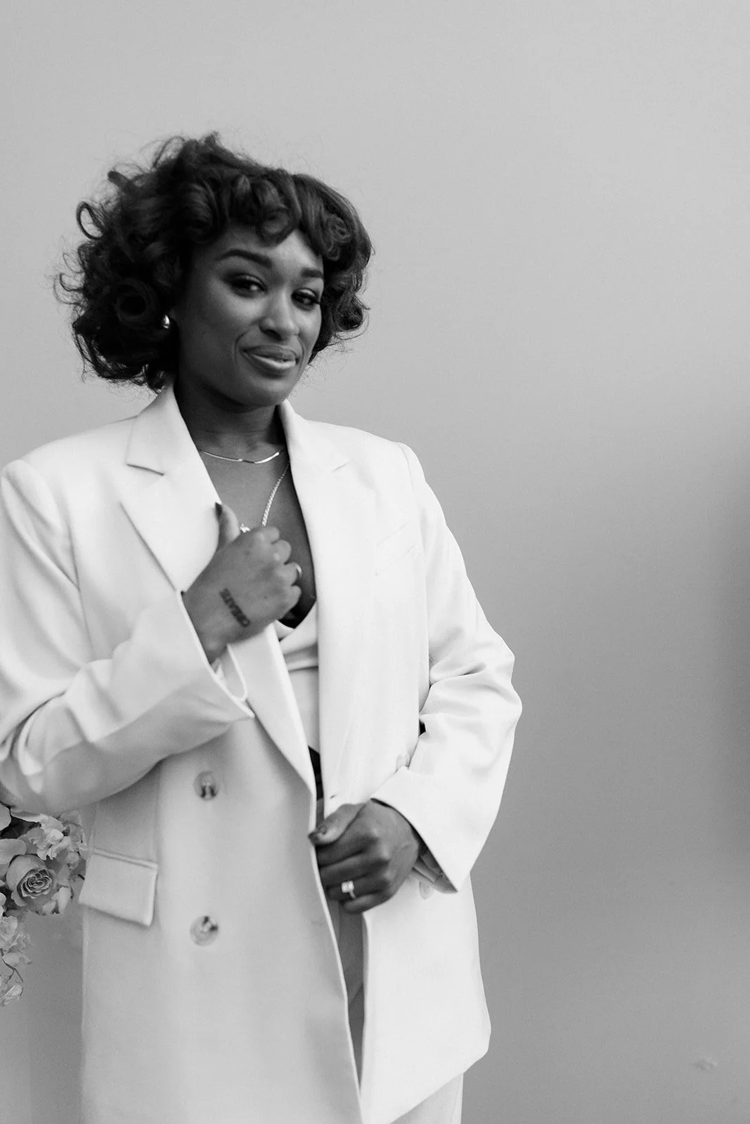 Black and white photo of a woman with curly hair wearing a white blazer, giving a thumbs up, standing against a plain background.