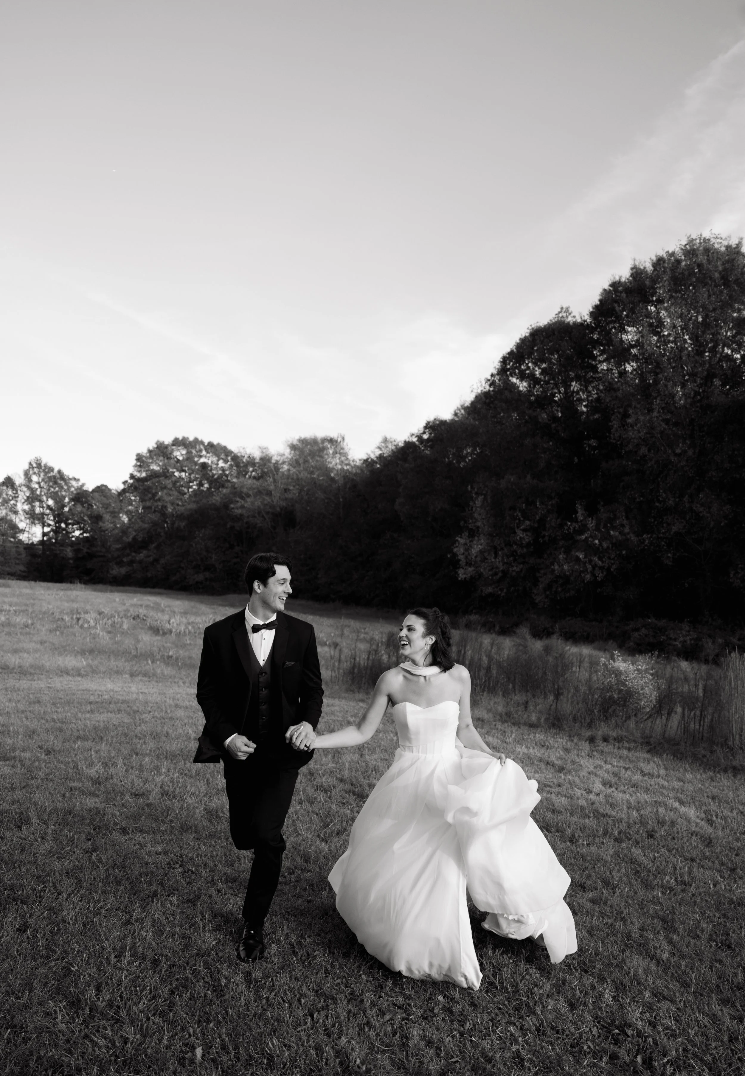 A black and white photograph of a smiling man and woman, dressed in formal wedding attire, walking hand in hand outdoors in a grassy field with trees in the background, during sunset or twilight.