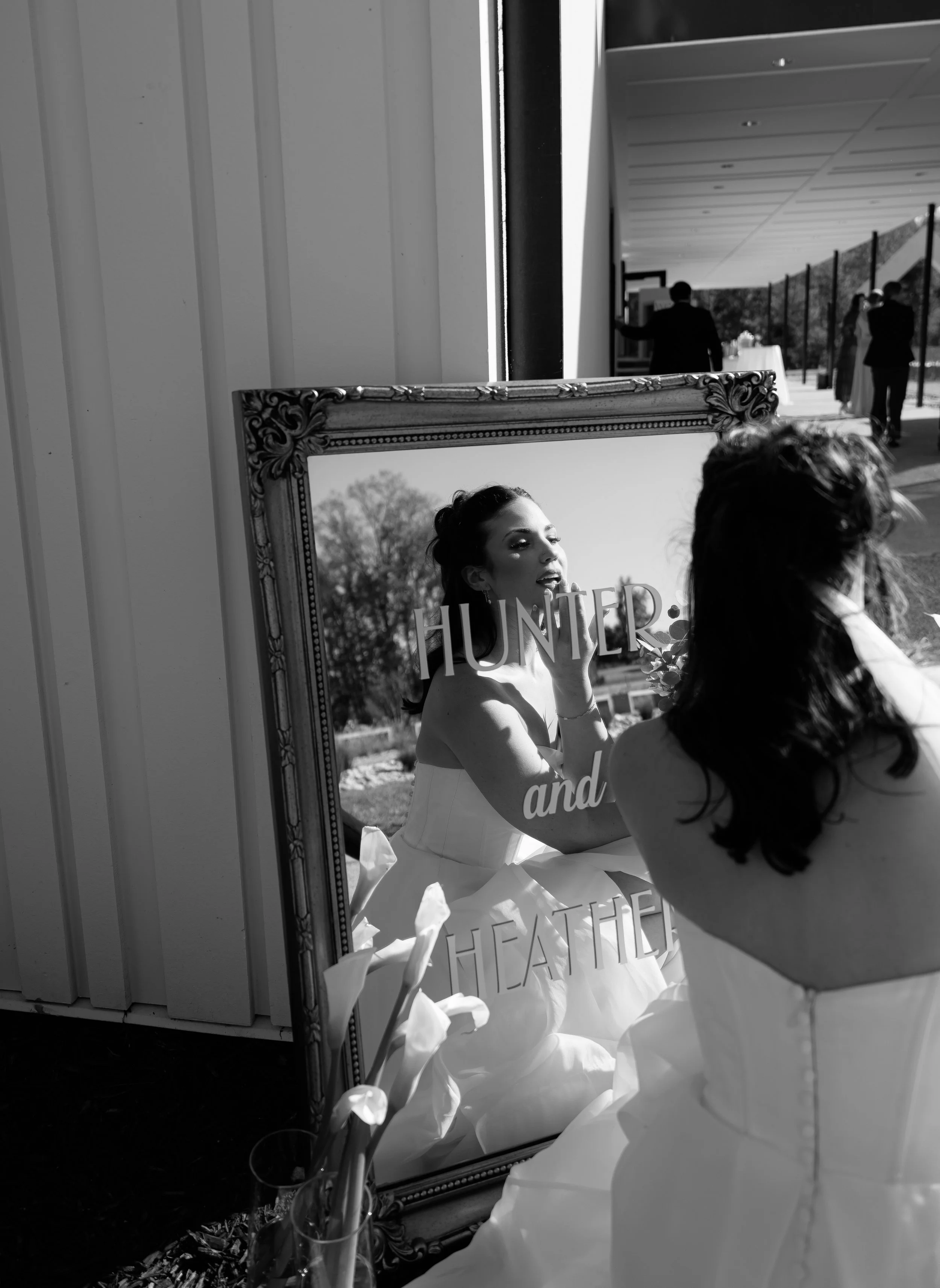 A woman in a wedding dress looks into a mirror, adjusting her makeup. Reflection shows her face with makeup, and the mirror has the words "HUNTER and HEATHER" on it. In the background, wedding guests are seen outdoors.