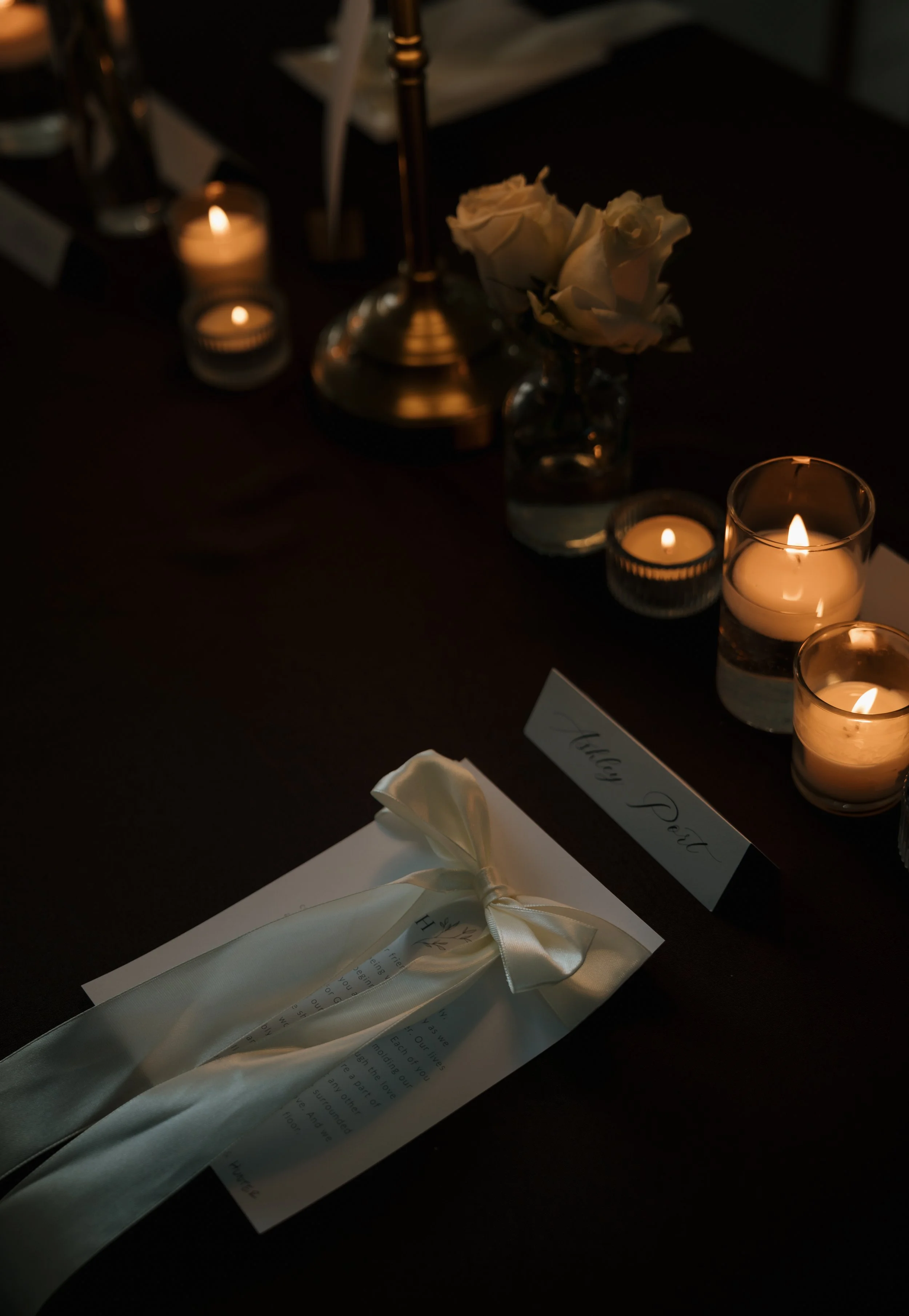 A table decorated with candles, white roses, and a folded white napkin tied with a ribbon. A name card with the text 'Yung P.' is visible on the table.