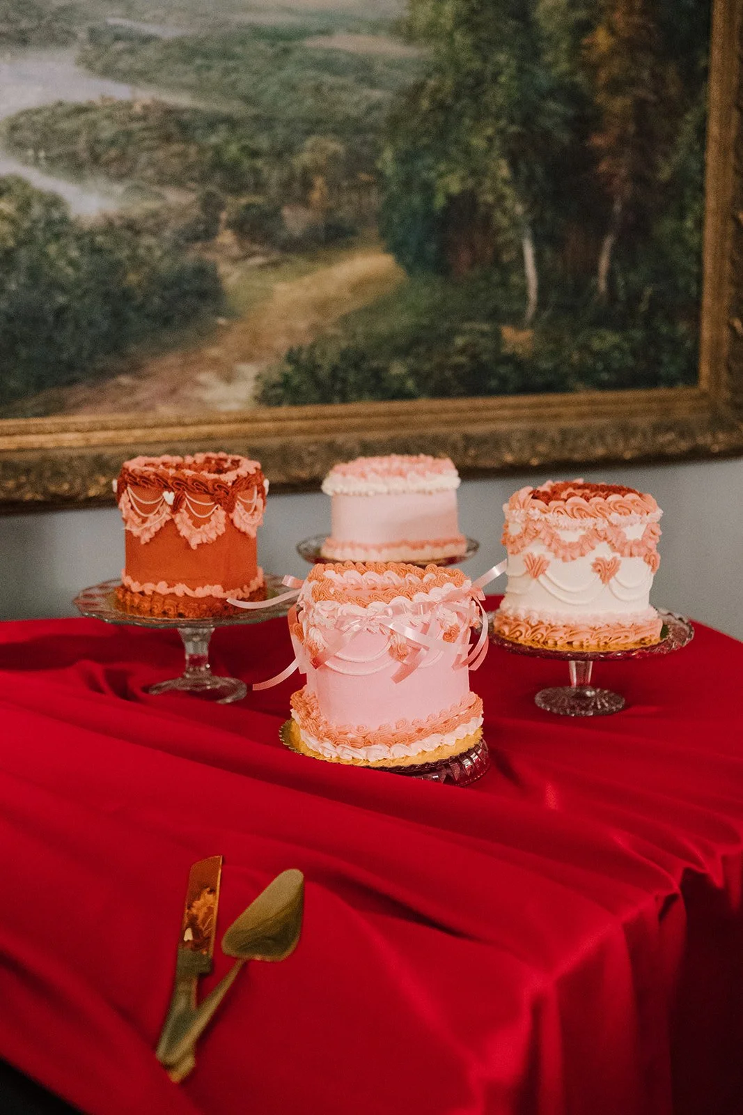Four pink and white decorated cakes on glass cake stands on a red tablecloth, with a gold-colored cake serving set in the foreground and a landscape painting hanging on the wall in the background.