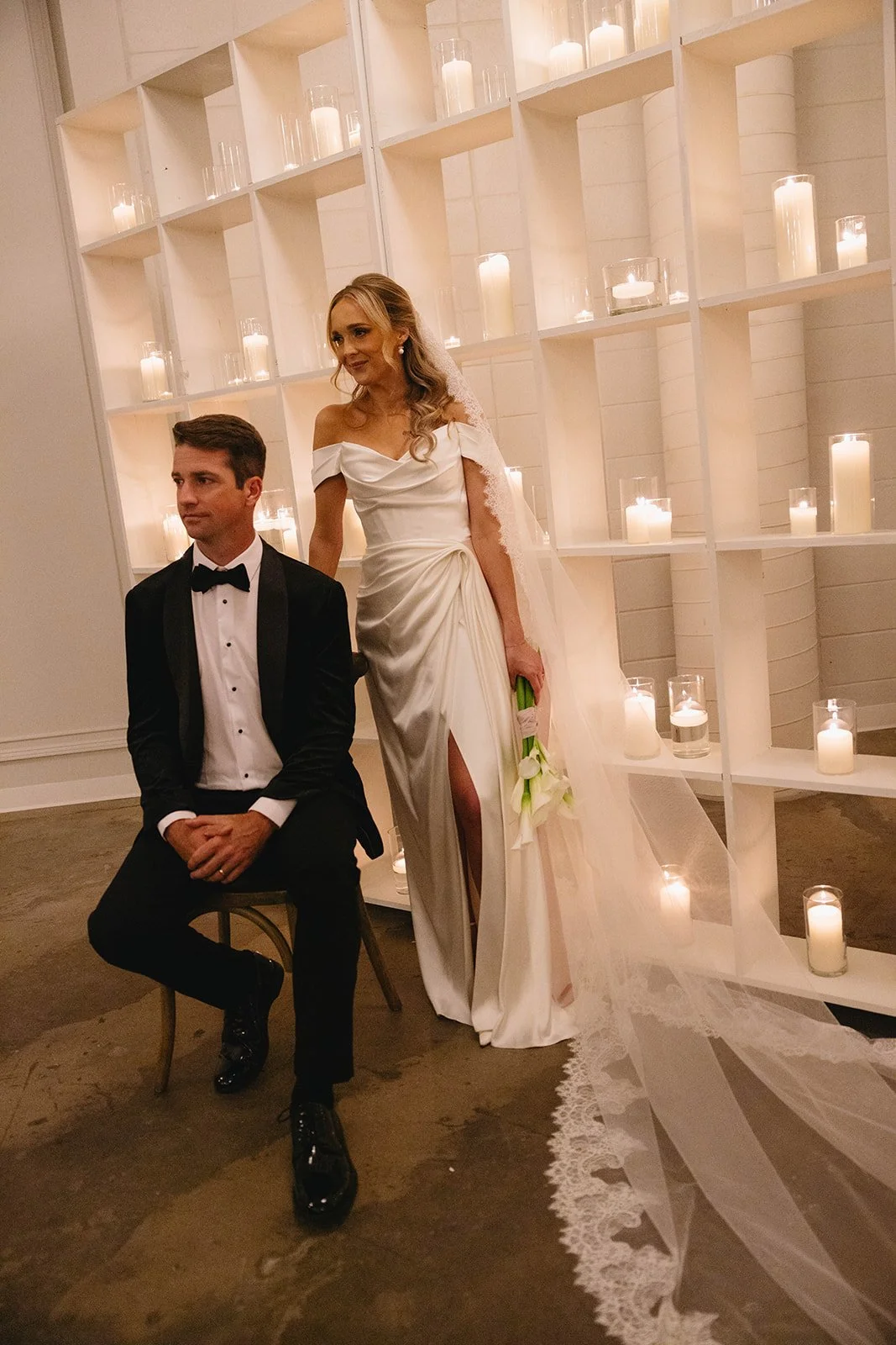 Bride and groom at their wedding with candles in the background. The bride is standing in a white wedding dress holding a bouquet, and the groom is sitting in a tuxedo.