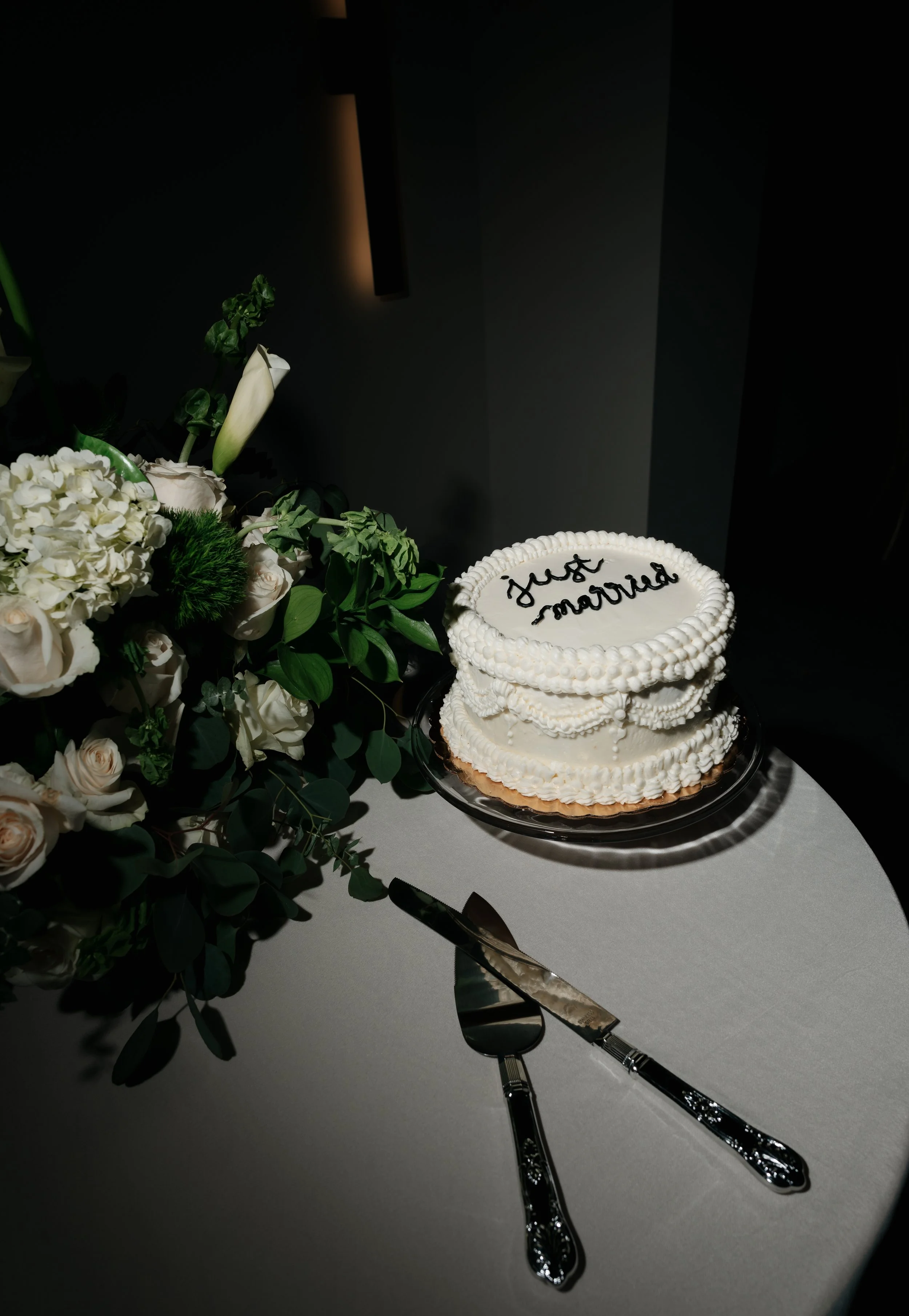A wedding cake with 'just married' written on top and a bouquet of white flowers on a table, with a cake knife and serving spatula in front.