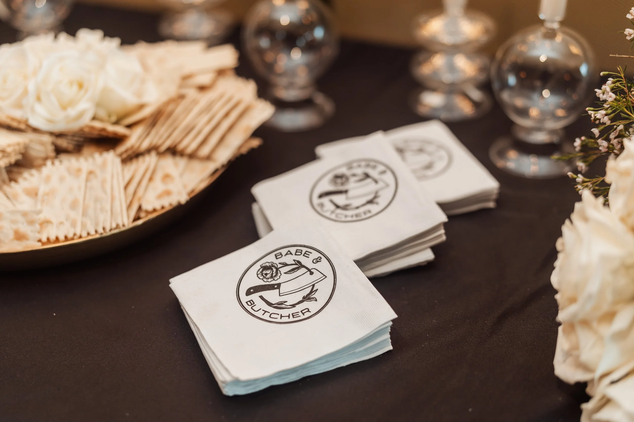 Stack of napkins with a logo that reads 'Babe & Butcher' next to a knife and rose, an array of glasses, and a plate of crackers and cheese on a dark table.