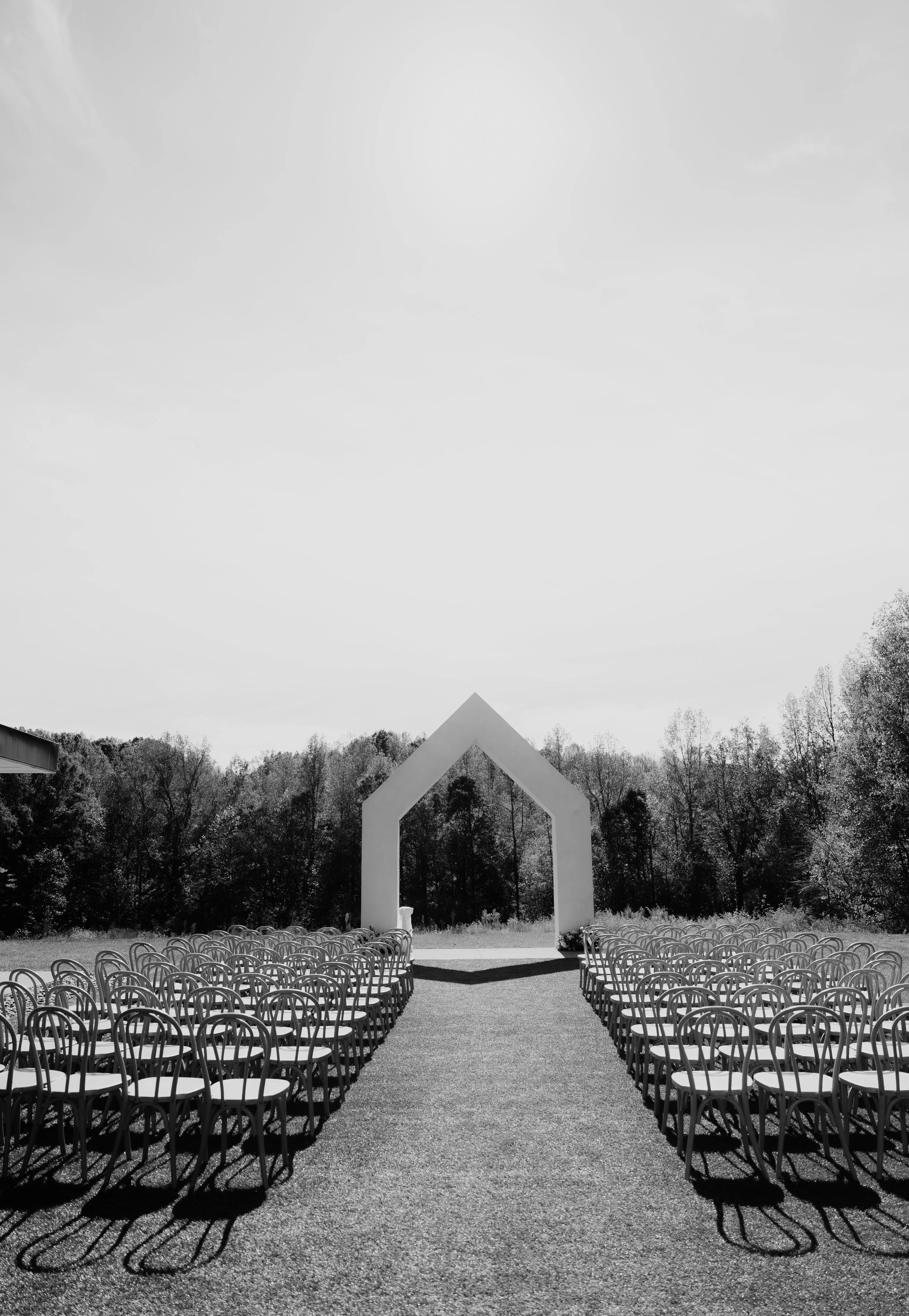 Outdoor wedding ceremony setup with chairs arranged in rows facing a large geometric arch and a backdrop of trees under a partly cloudy sky.