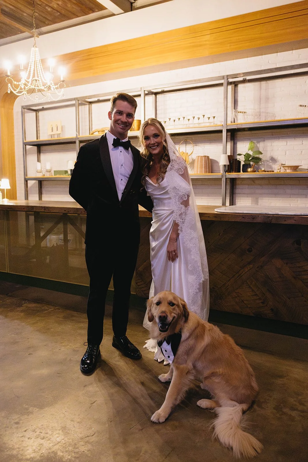 A bride and groom dressed in wedding attire standing in a modern, rustic venue with a golden retriever sitting in front of them wearing a tuxedo bowtie. The bride is wearing a lace wedding gown with a veil, and the groom is wearing a black tuxedo wit
