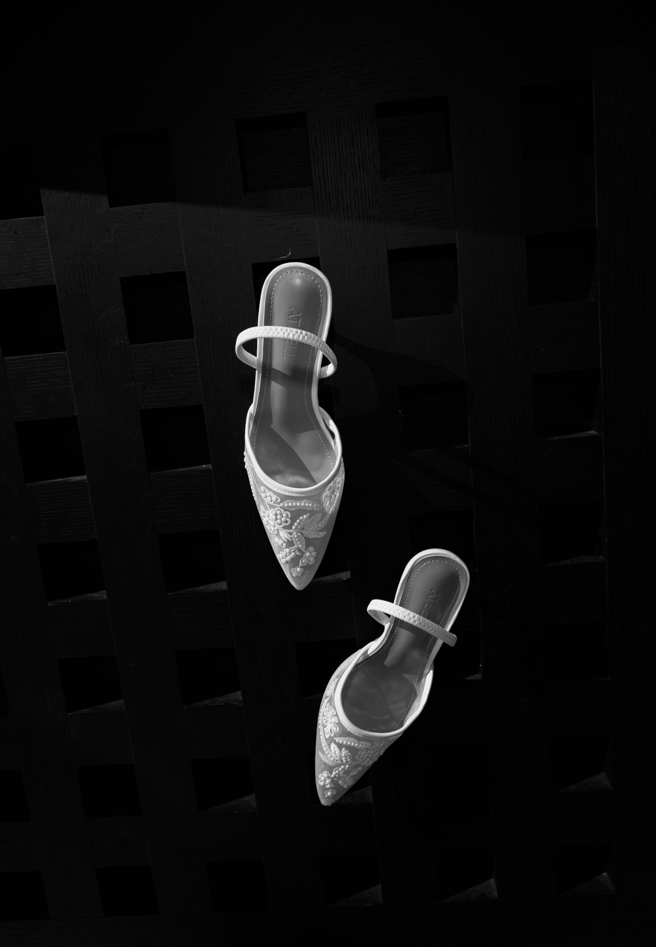 Two white high-heeled shoes with embroidery on a dark wooden grid surface, with sunlight casting shadows.