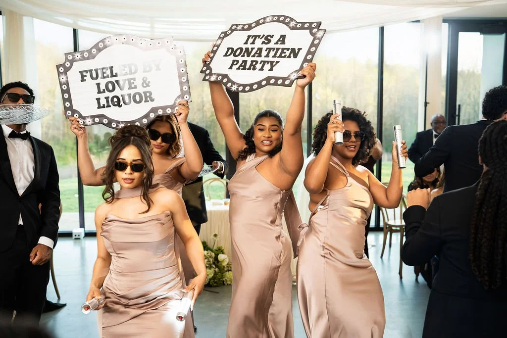 Group of women in satin dresses holding signs at a celebration event, with one sign reading 'Fuelled by Love & Liquor' and another 'It's a Donatien Party', in a room with large windows and outdoor greenery.