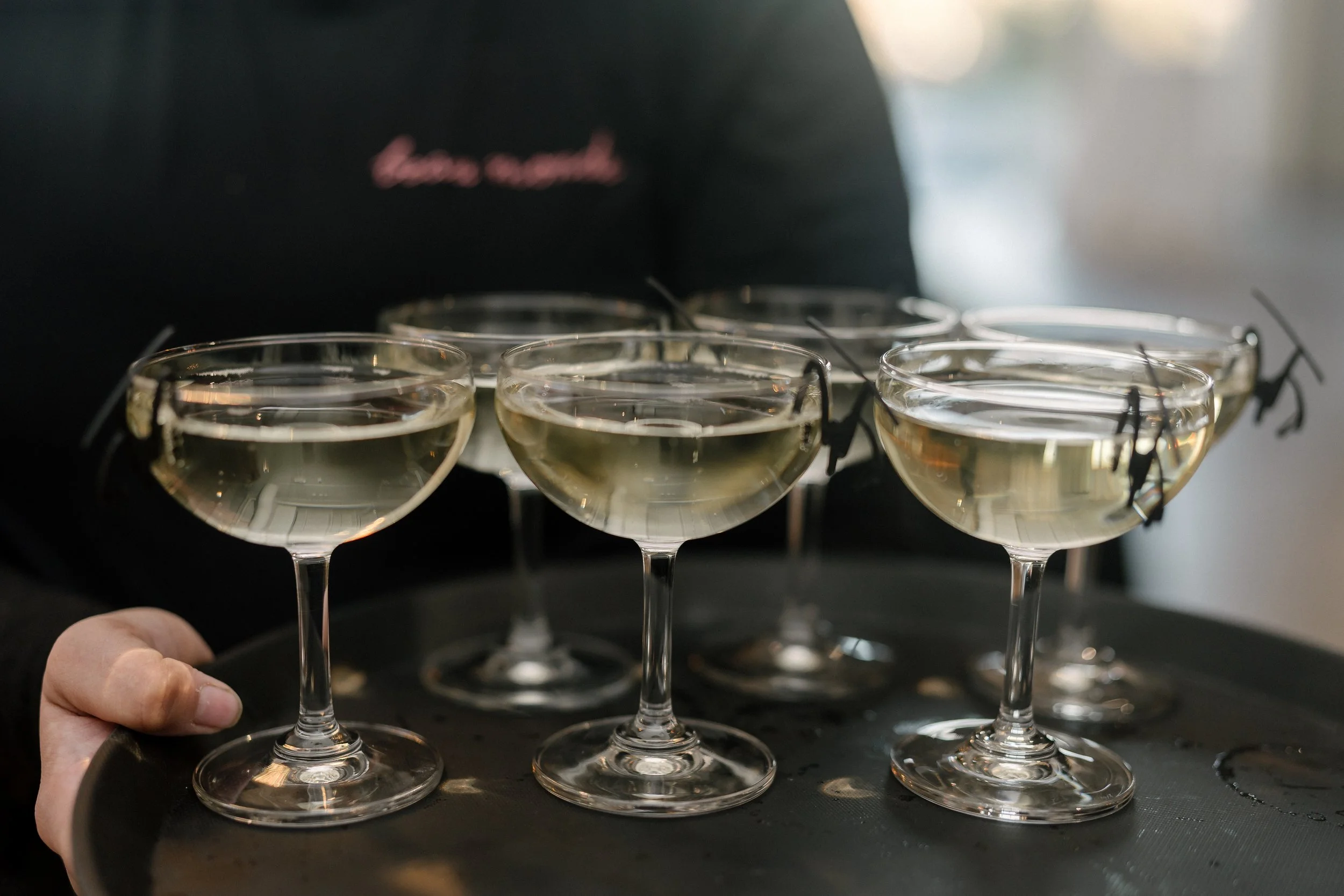 A person holding a tray with six champagne glasses filled with a light-colored sparkling drink.