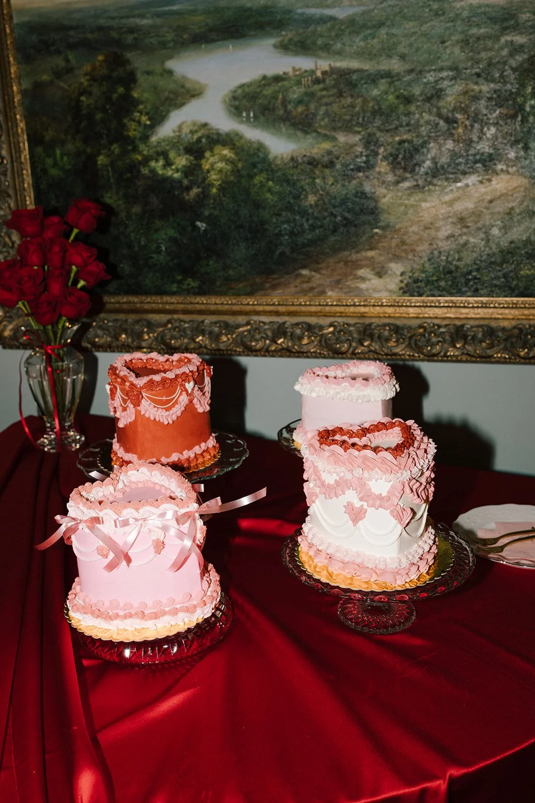 Three pink layered cakes decorated with pink and white icing, topped with hearts and ribbons, on a red tablecloth. A bouquet of red roses in a glass vase is on the left, and a landscape painting hangs on the wall behind.