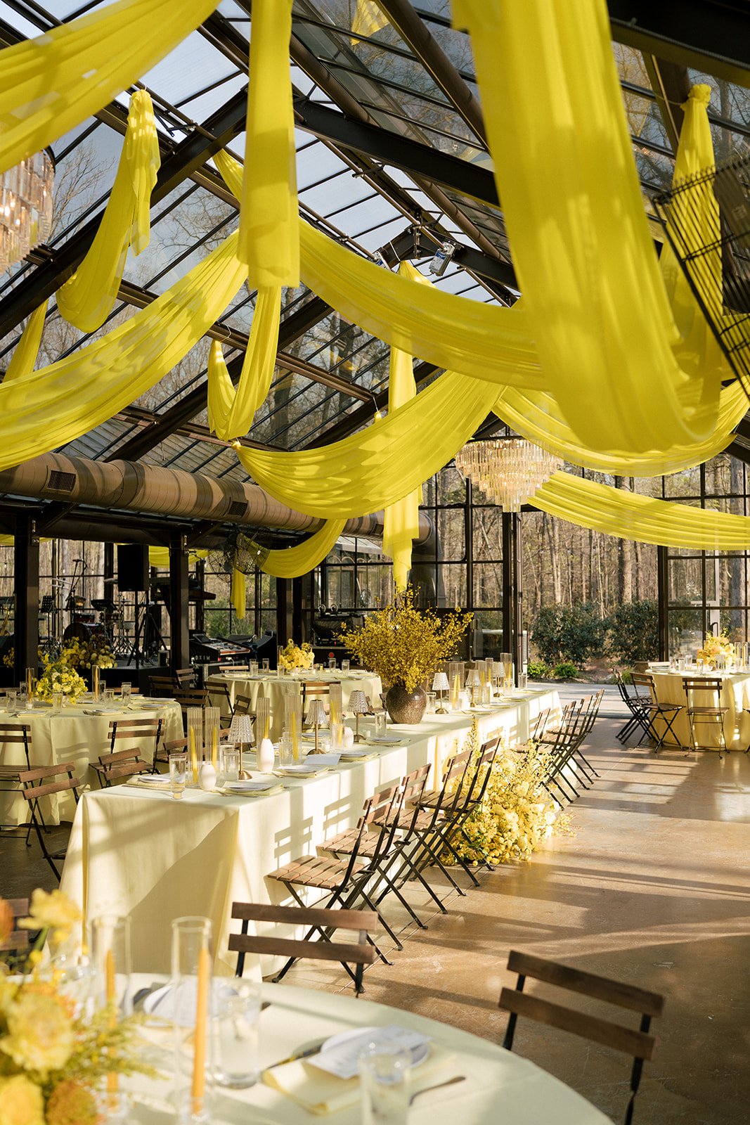 Wedding reception setup with yellow drapery, floral arrangements, and tables inside a glass conservatory.