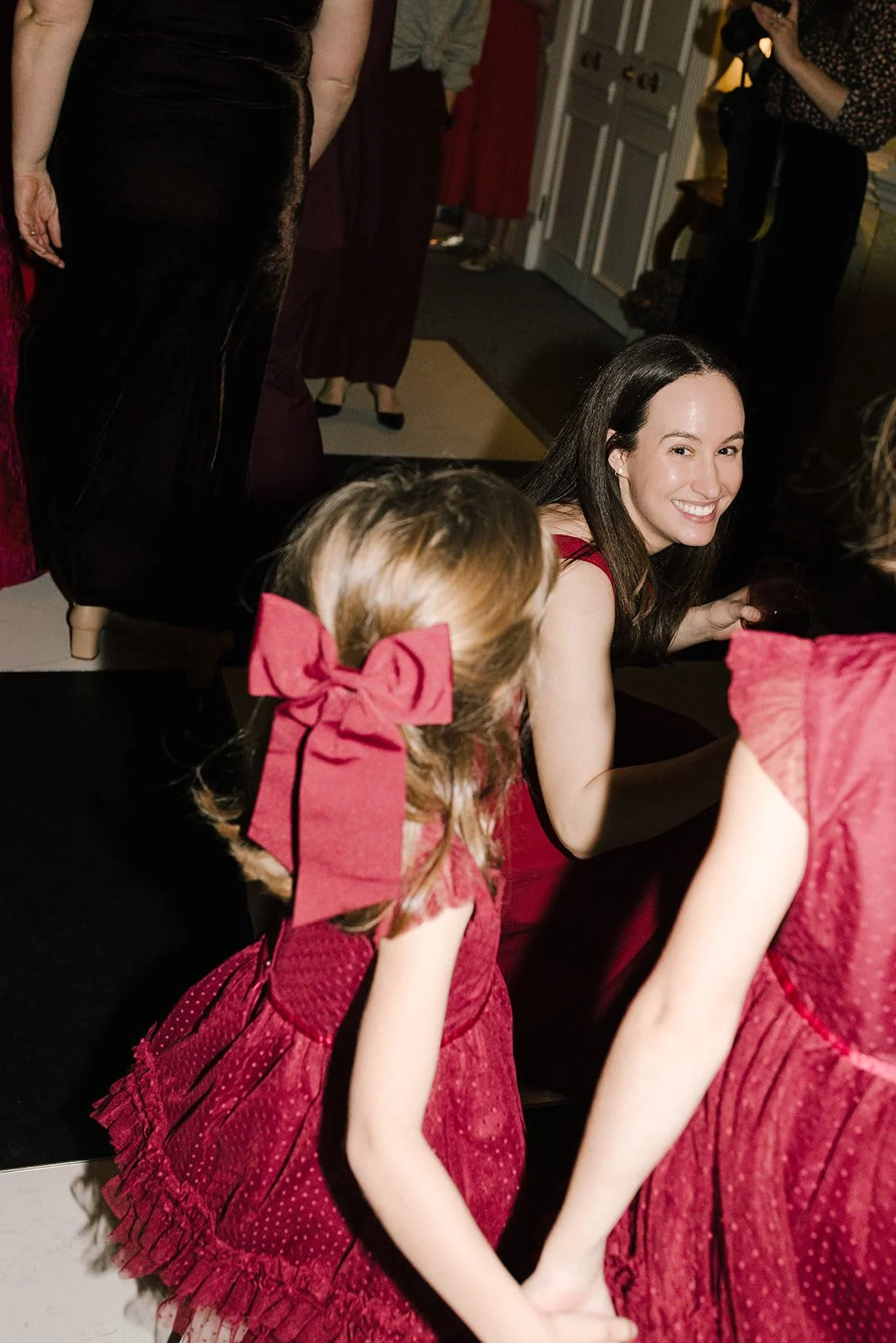A smiling woman in a red dress sitting on the floor at a social gathering, surrounded by children in matching red dresses with bows, with other people in the background.