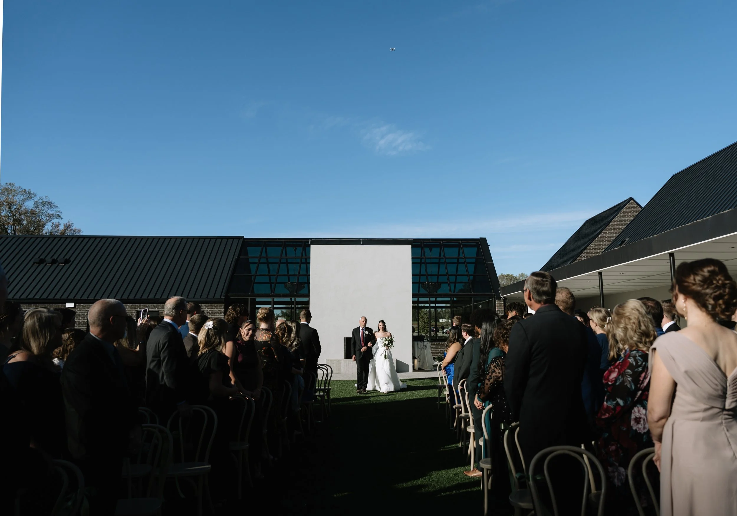 Outdoor wedding ceremony with guests seated, bride and groom standing in front of a white altar under a clear blue sky.