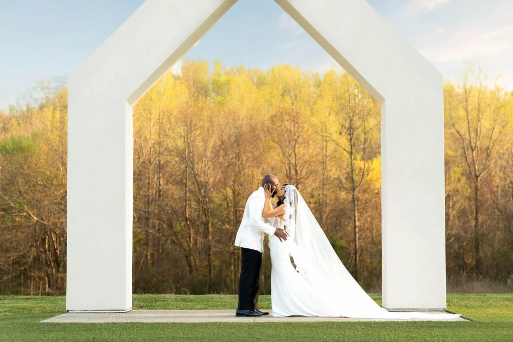 A couple, a bride in a white wedding gown and a groom in a white jacket, embracing and kissing under a white, house-shaped structure outdoors during fall, with trees showing yellow and orange leaves in the background.