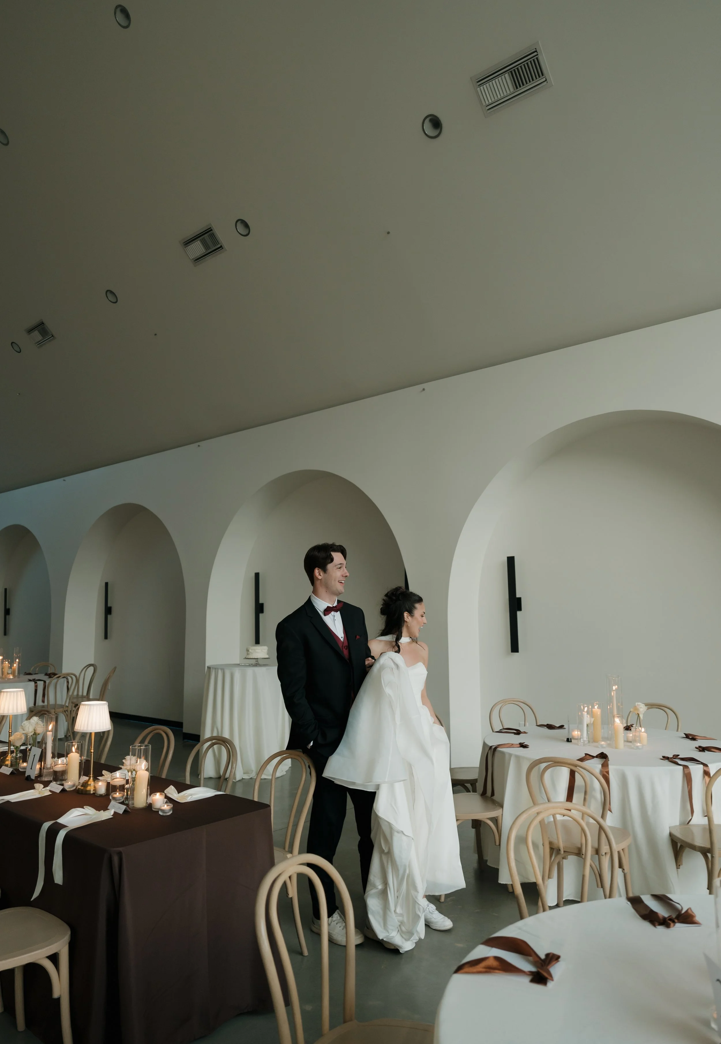 A bride and groom standing in a decorated reception hall with round tables, candles, and white walls with arches.