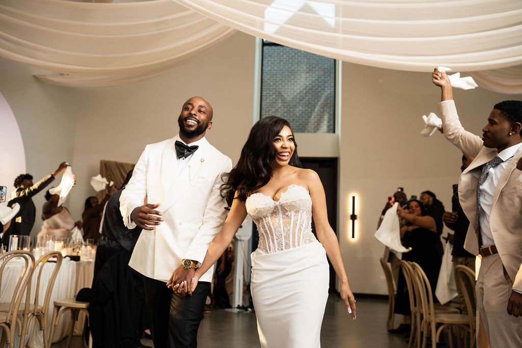 Bride and groom holding hands and smiling at their wedding reception, surrounded by guests celebrating.