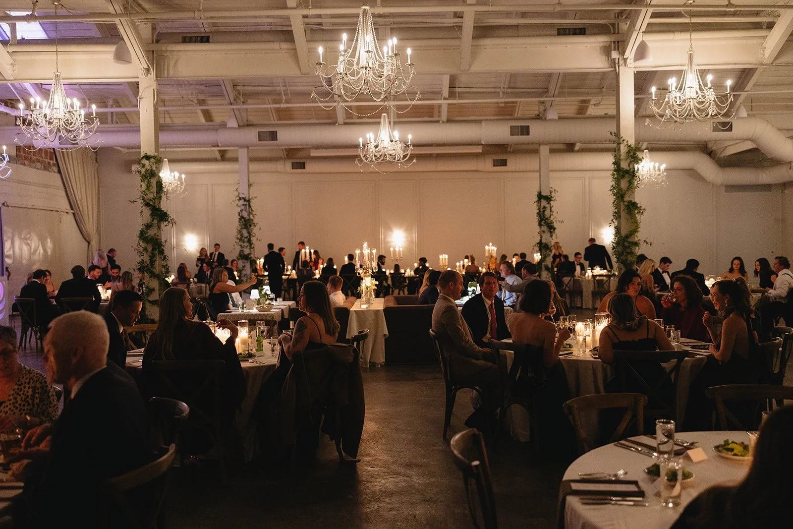 A large indoor wedding reception with guests seated at round tables decorated with candles and flowers, chandeliers hanging from the high ceiling, and waitstaff serving food in the background.