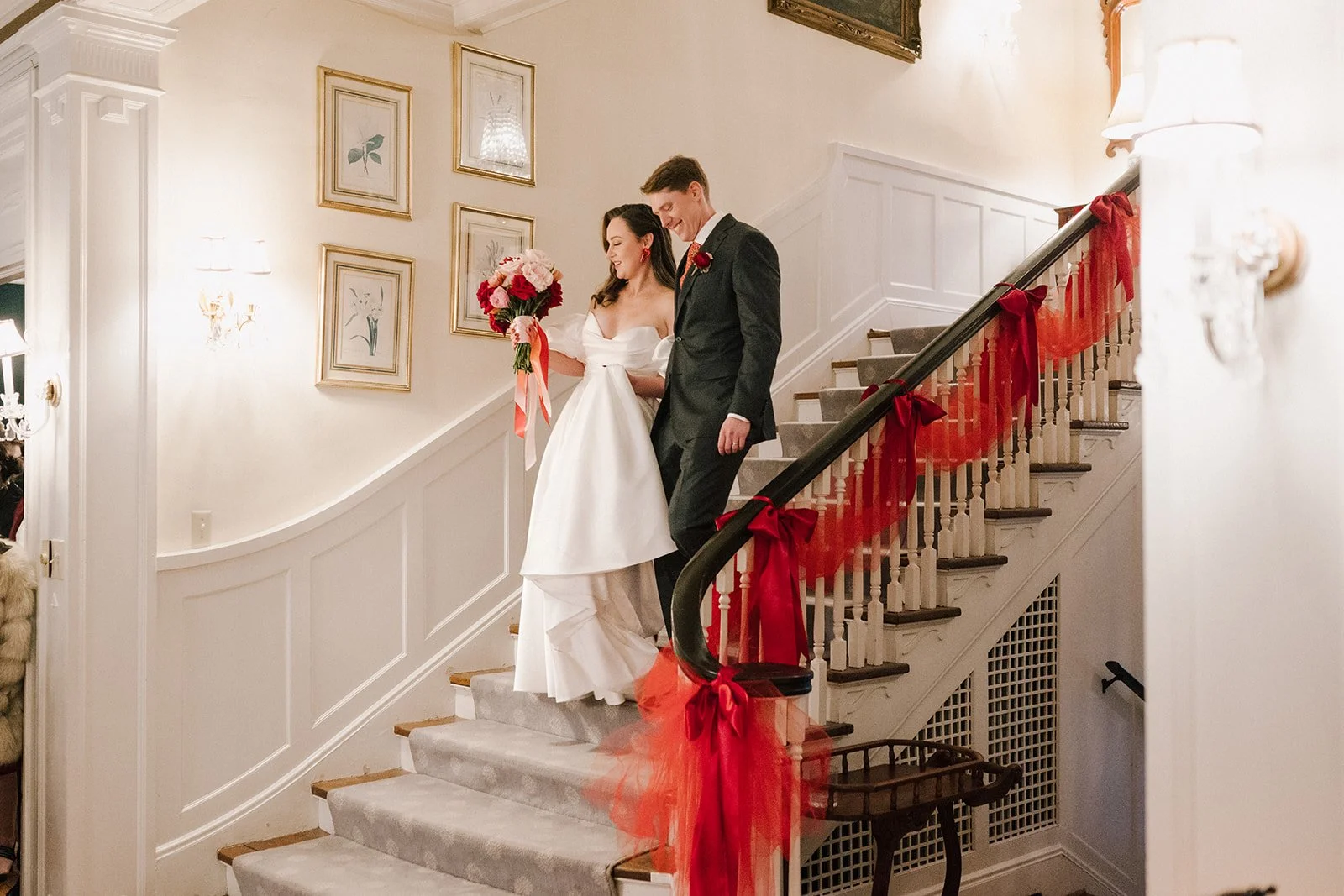 A bride in a white wedding gown and a groom in a dark suit standing on a staircase decorated with red ribbons and bows, with framed botanical art on the wall behind them.
