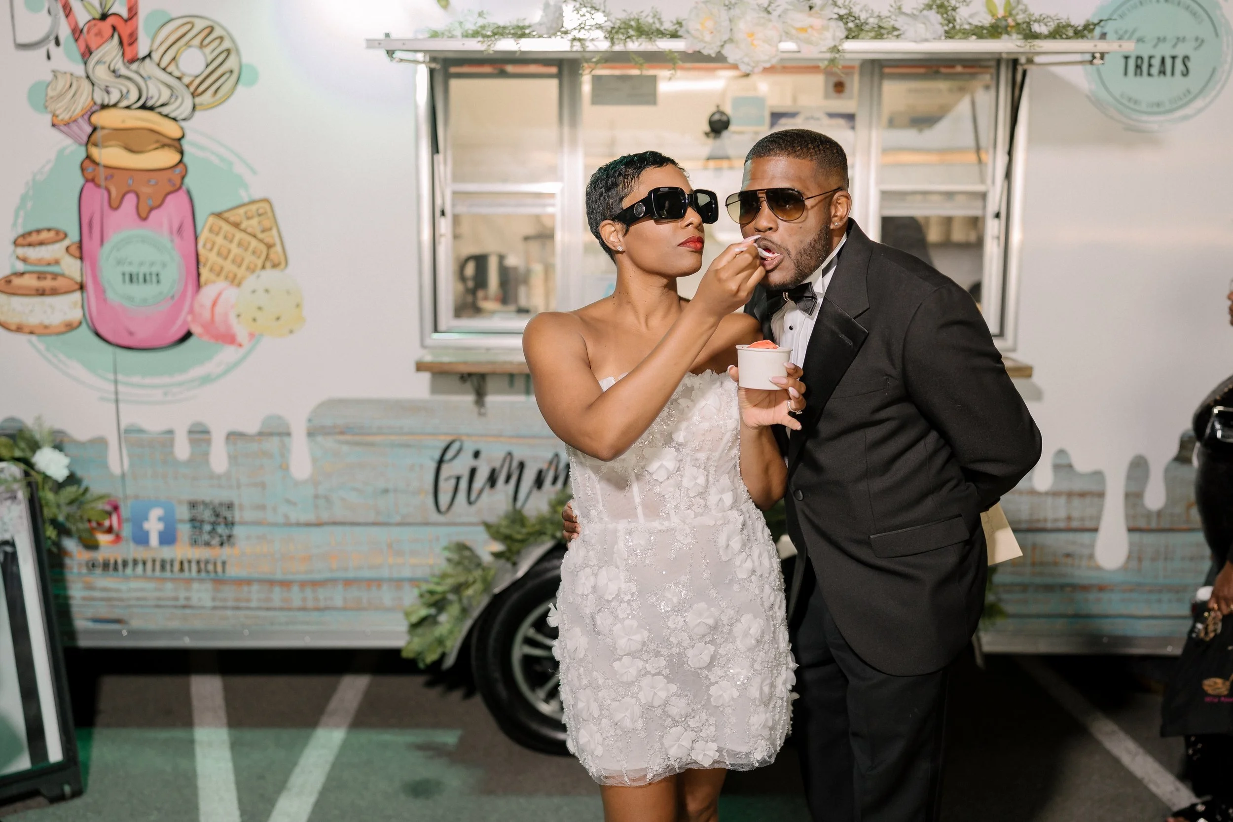 A bride and groom in wedding attire sharing a moment at an outdoor celebration, with a dessert truck in the background.