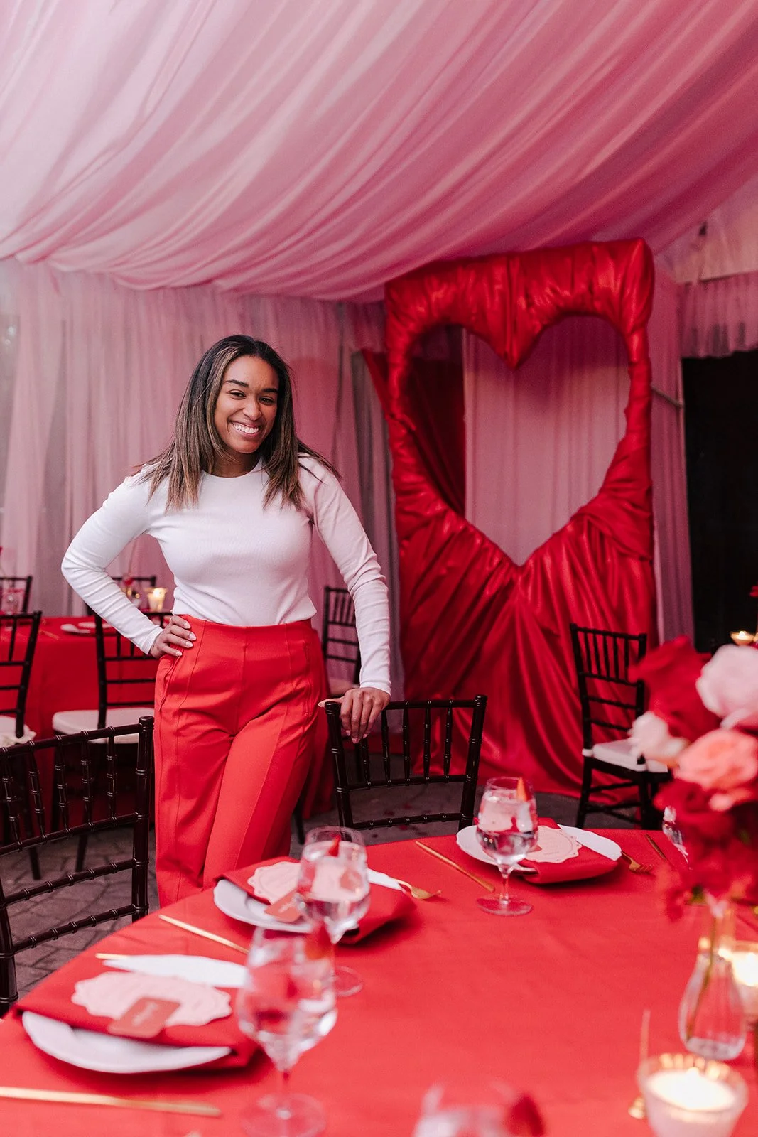 A woman standing in a decorated event space with pink drapery and a red heart-shaped photo backdrop, wearing a white top and red pants, smiling at the camera.
