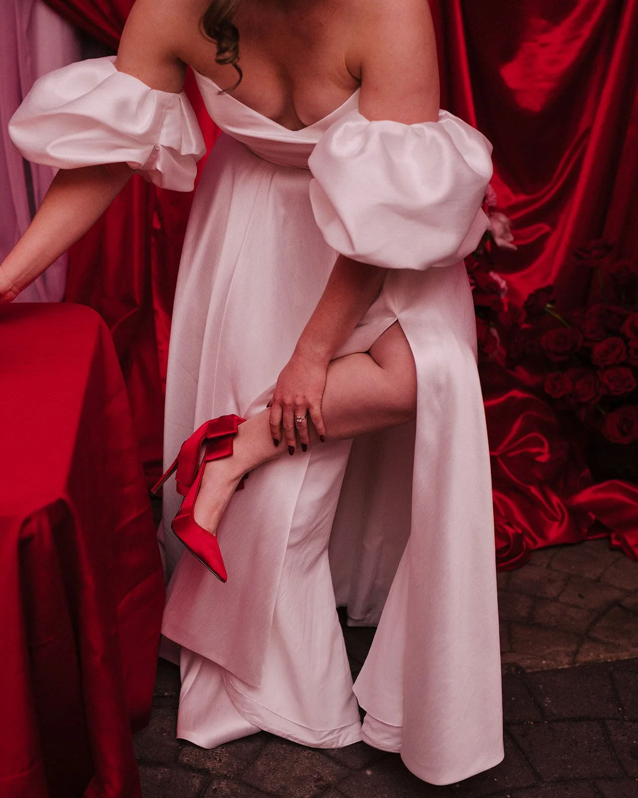 A woman is dressed in a white gown with large puffy sleeves, lifting her leg to show a red high heel shoe, against a background of red drapery and roses.