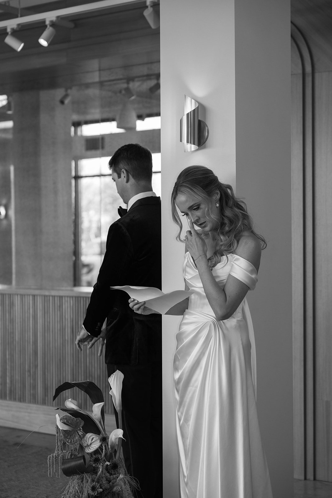 A bride in a white satin wedding dress is reading a letter with a distressed expression, standing next to a groom in a black tuxedo, who is looking down, both leaning against a corner in a modern indoor setting.