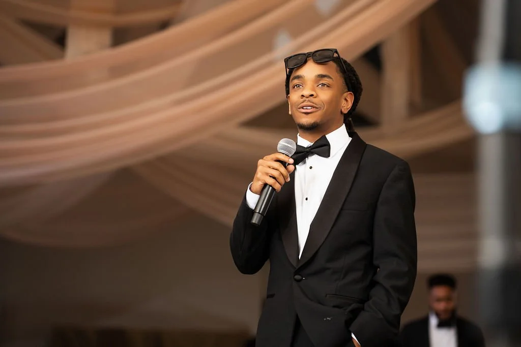 A young man in a black tuxedo with a bow tie, holding a microphone, standing in front of a draped fabric background at an event.