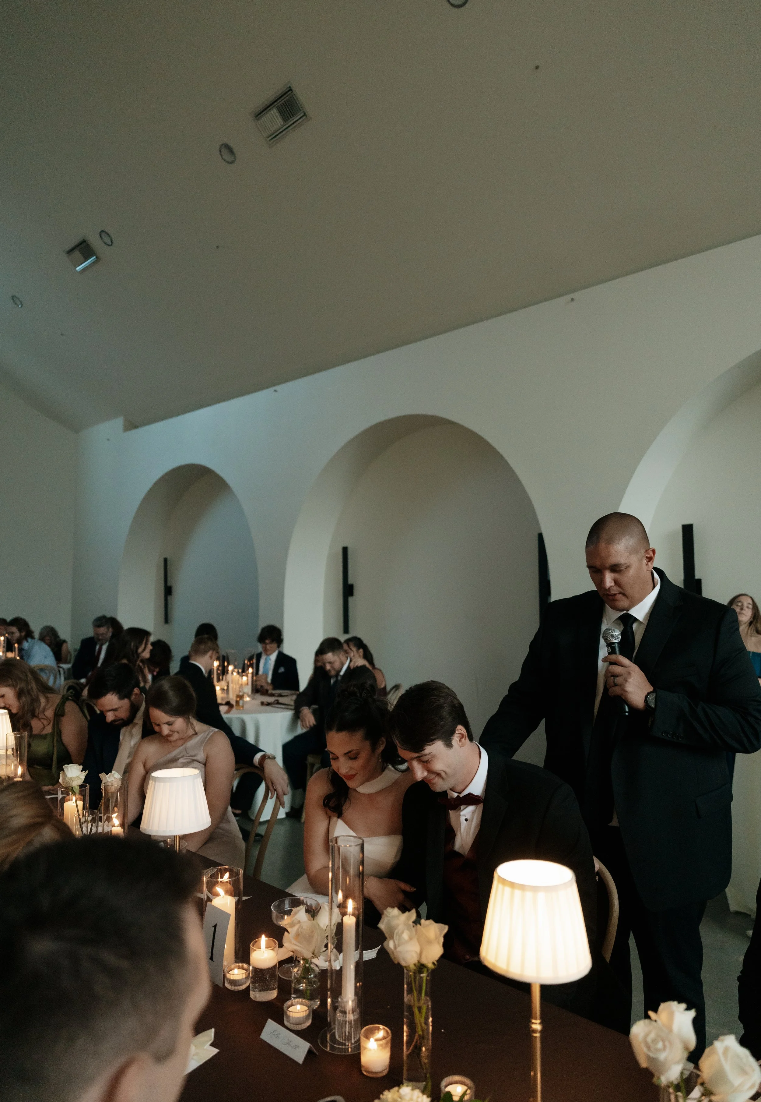 A wedding reception scene with guests seated at tables decorated with candles, flowers, and tableware, while a man in a suit presents or speaks, and other guests listen or interact in a room with white walls, arched doorways, and ceiling lights.