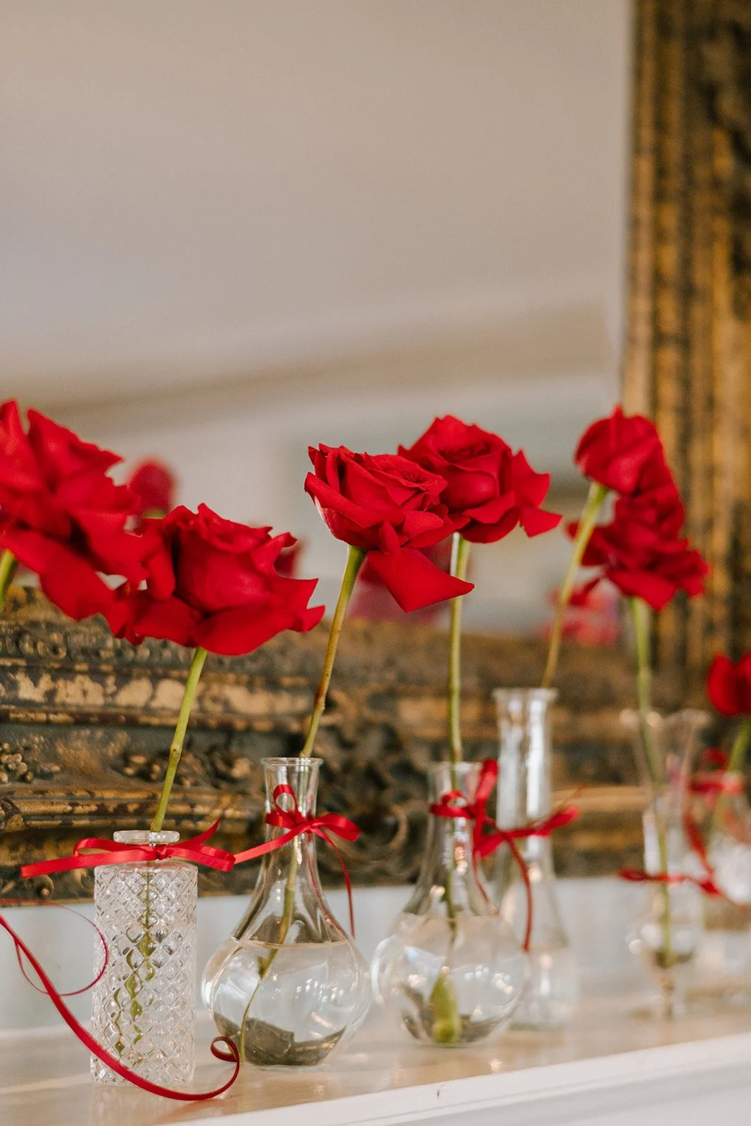 Red roses in glass vases with red ribbons on a table, with a blurred ornate gold-framed mirror or picture frame in the background.