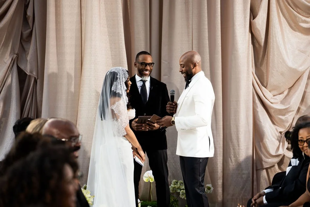 A couple getting married, standing in front of a beige curtain, with a man officiating the ceremony. The bride wears a traditional wedding dress and veil, and the groom is in a black suit. Guests are seated around them.