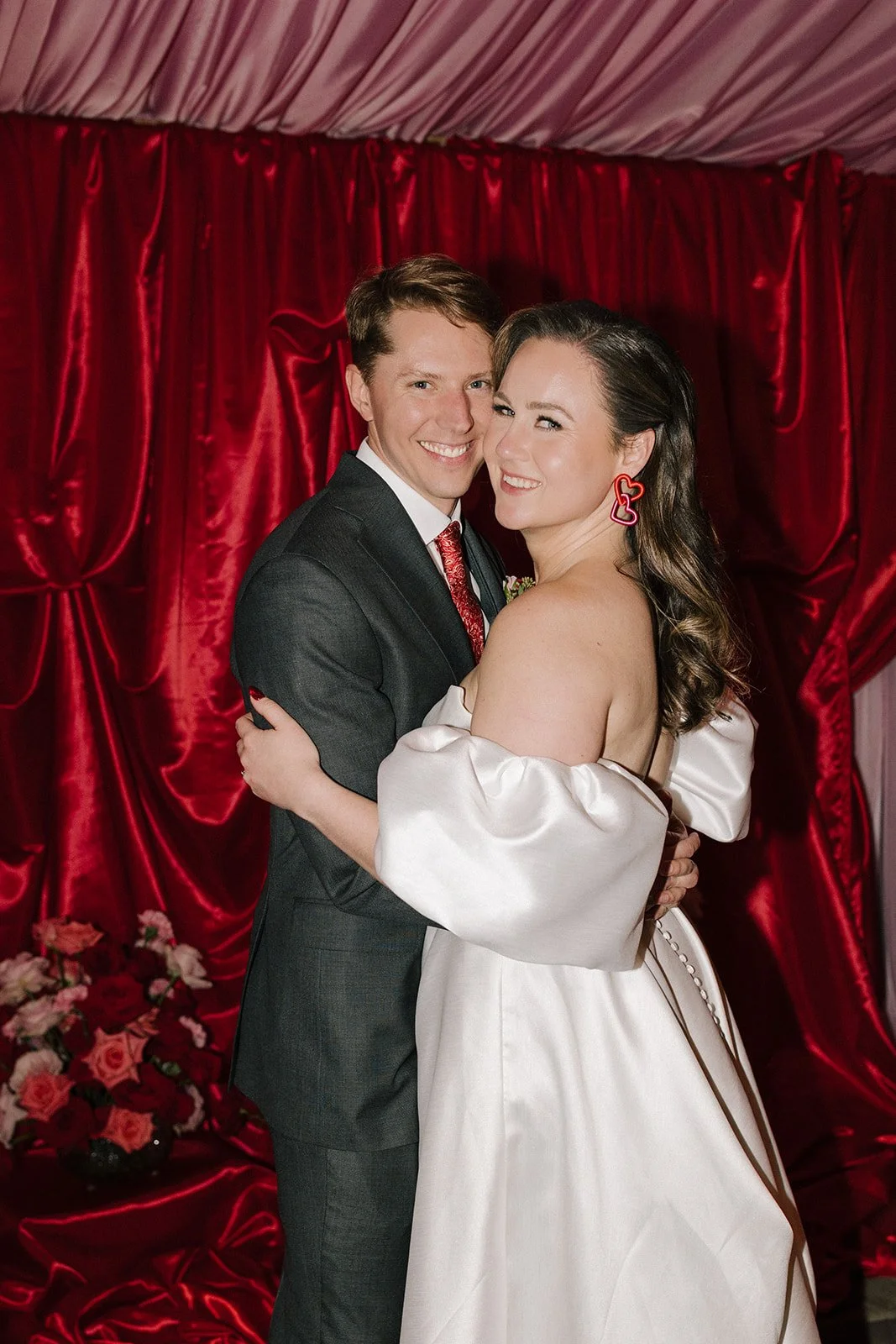 A happy couple in wedding attire embracing, standing in front of a red velvet curtain with a floral arrangement in front.
