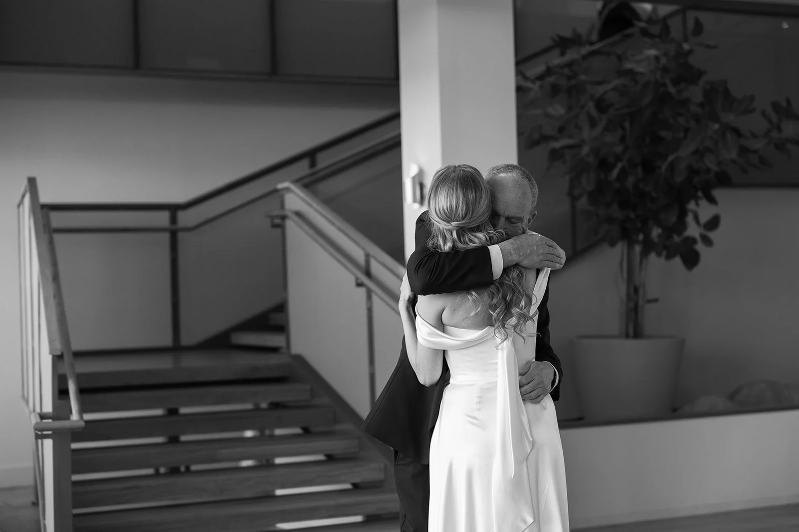 A bride and groom sharing a heartfelt hug during their wedding inside a modern venue with stairs and a potted plant in the background, all in black and white.